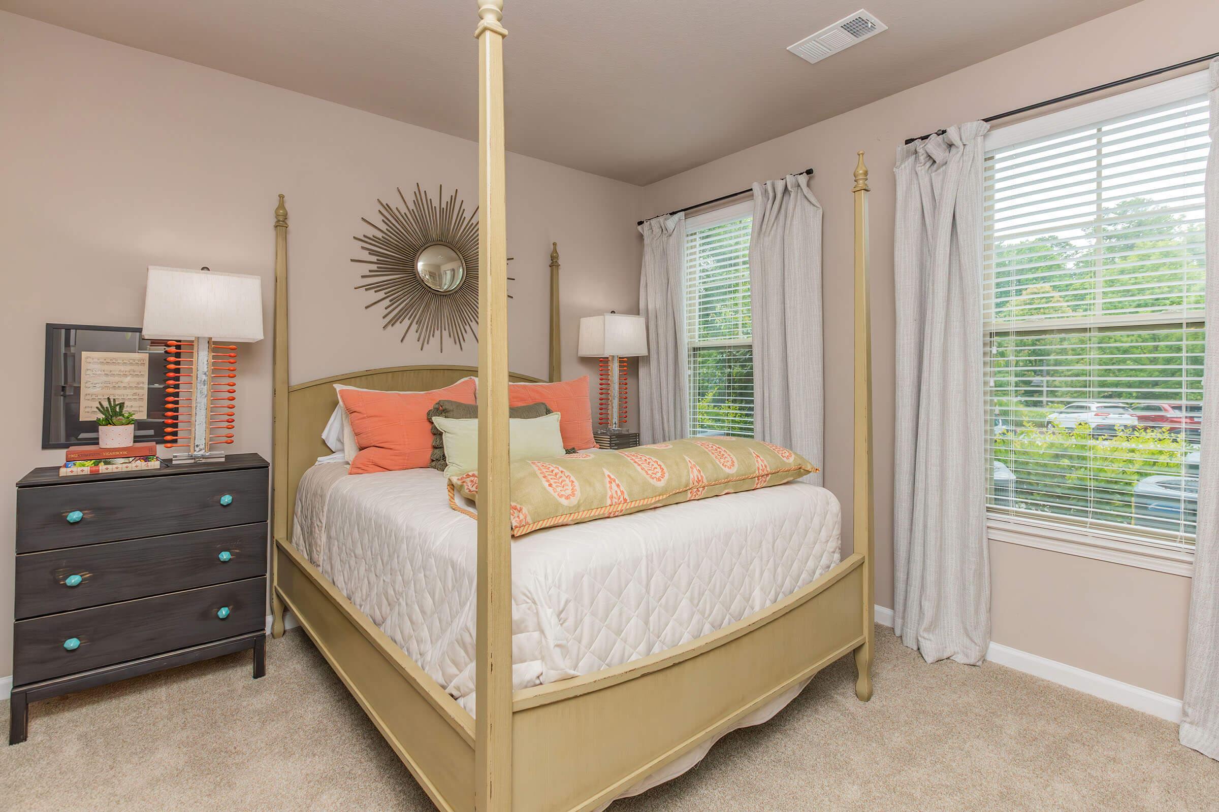 A cozy bedroom featuring a four-poster bed with an orange and green decorative pillow, flanked by two lamps on nightstands. The walls are painted a soft beige, and large windows with sheer curtains allow natural light to brighten the room. A dark wood dresser is visible in the corner.