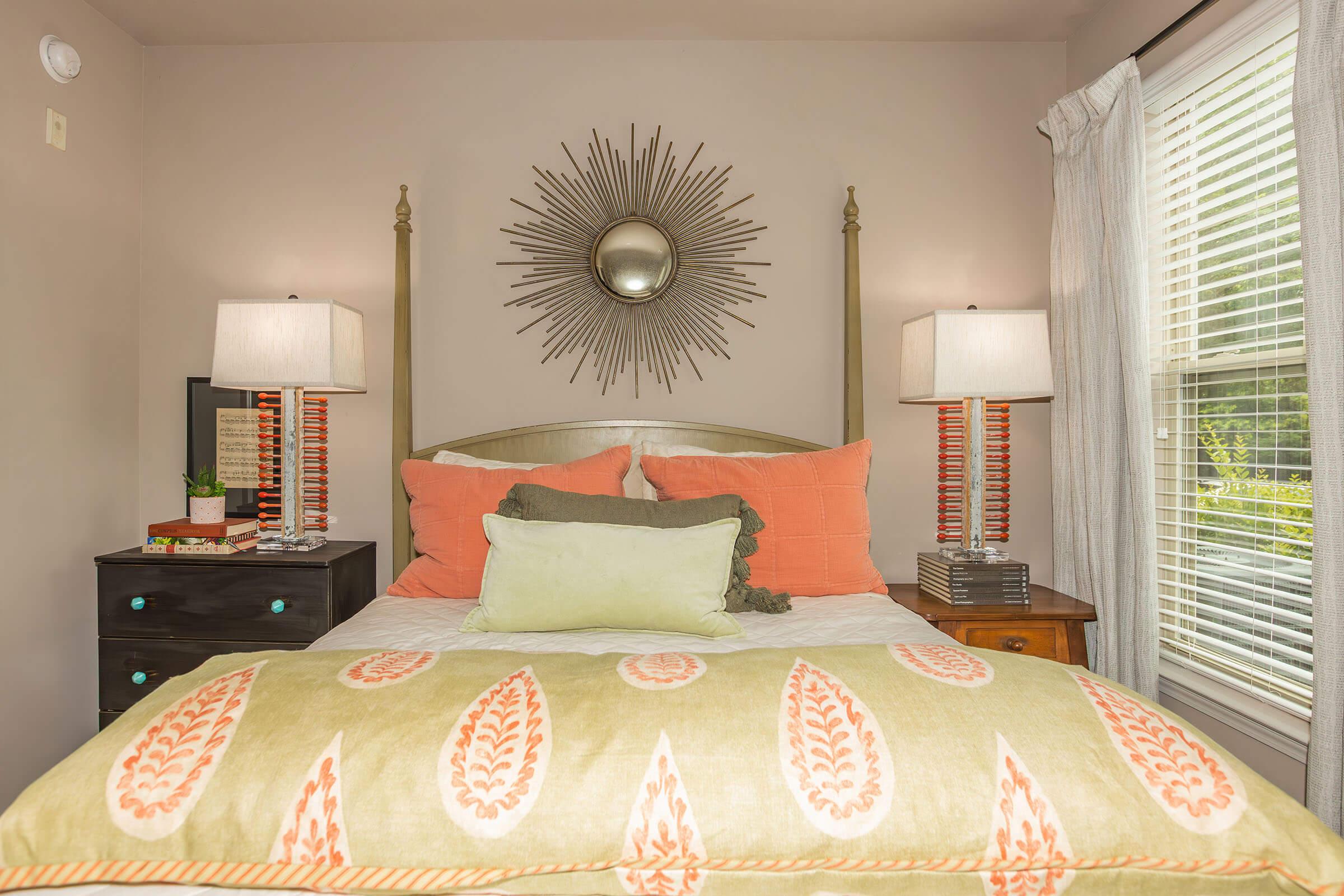 A cozy bedroom featuring a bed with orange and green pillows, a decorative blanket with a leaf pattern, two bedside lamps with striped bases, and a sunburst mirror above the headboard. Shelves hold books and a small plant, while natural light filters through white curtains.