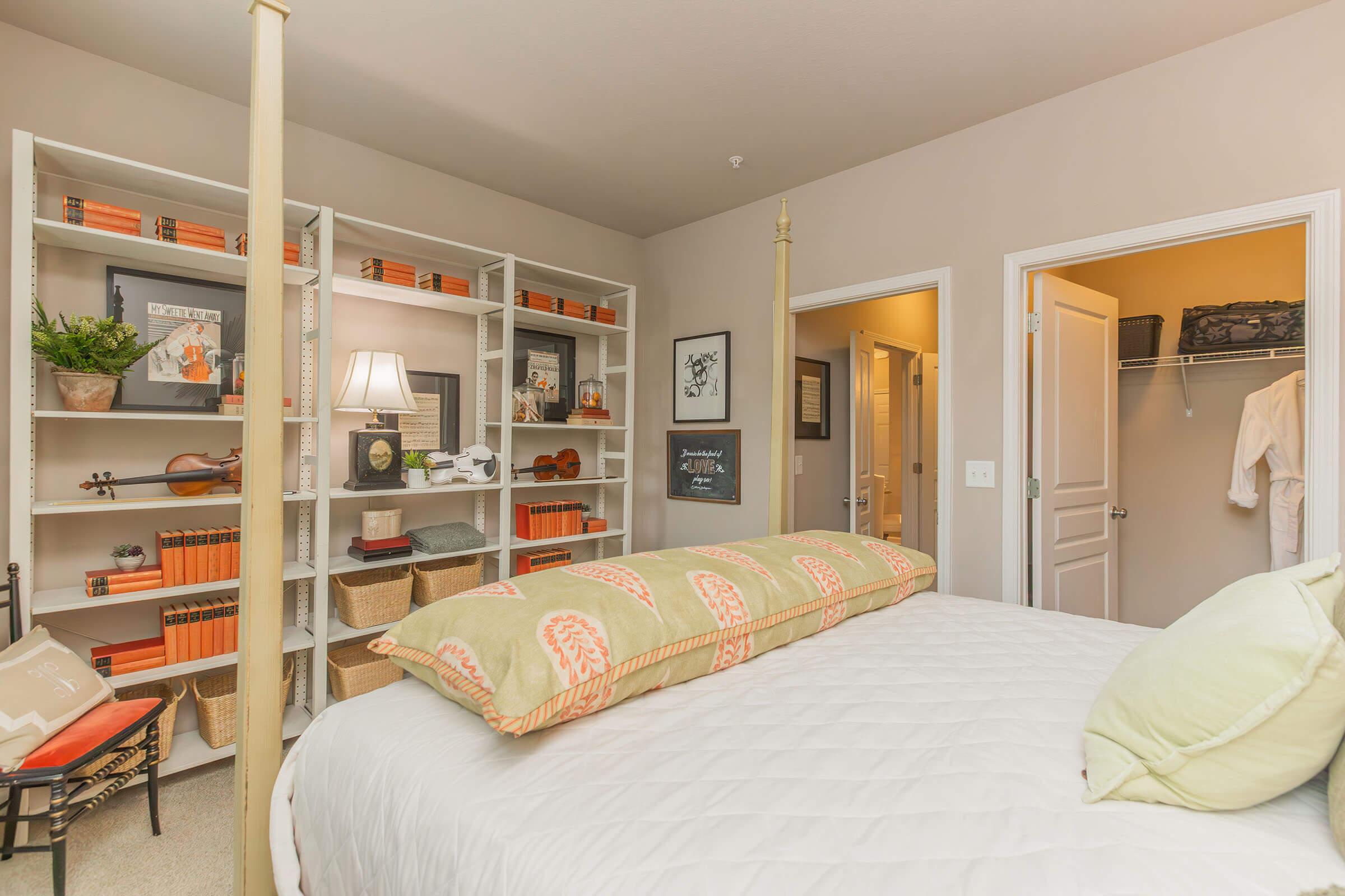 A cozy bedroom featuring a decorative bed with a long patterned pillow, bookshelves filled with books and decorative items, a lamp providing soft lighting, and two doors leading to adjoining rooms. The warm, neutral colors create a welcoming atmosphere.