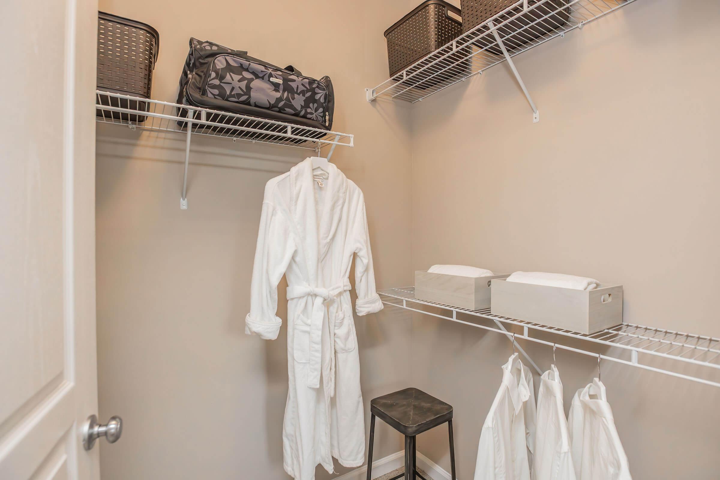A small storage space featuring white shelves with neatly organized baskets and boxes. A white robe hangs on a hook, while additional white robes are draped on a lower shelf. A simple black stool is positioned nearby, adding functionality to the cozy, well-organized area.