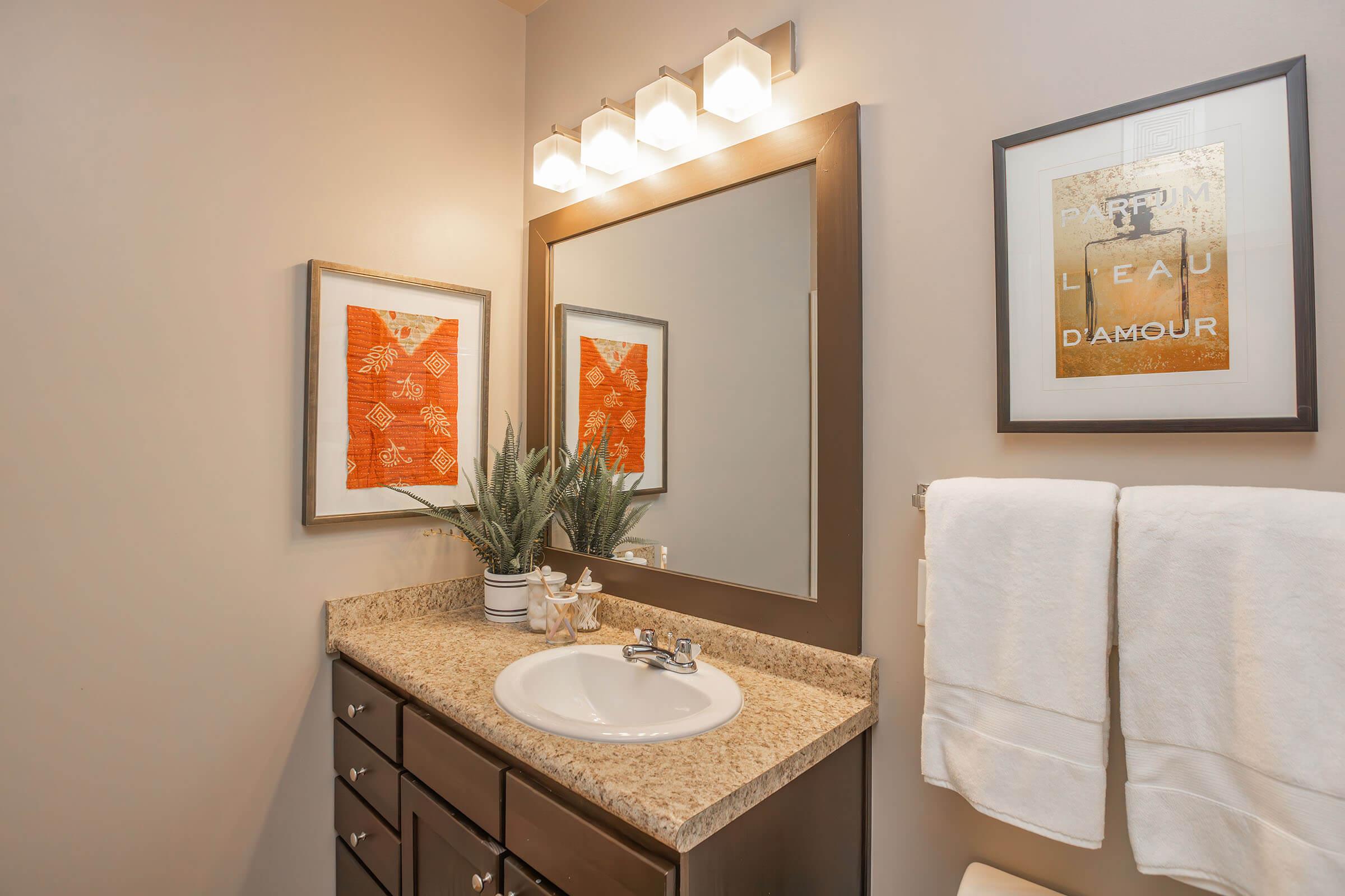 A modern bathroom featuring a granite countertop with a sink, decorative items, and a small plant. Above the sink, there is a large mirror and three light fixtures. The wall has a framed artwork with an orange design. Two white towels hang on a rack nearby, adding to the overall decor.