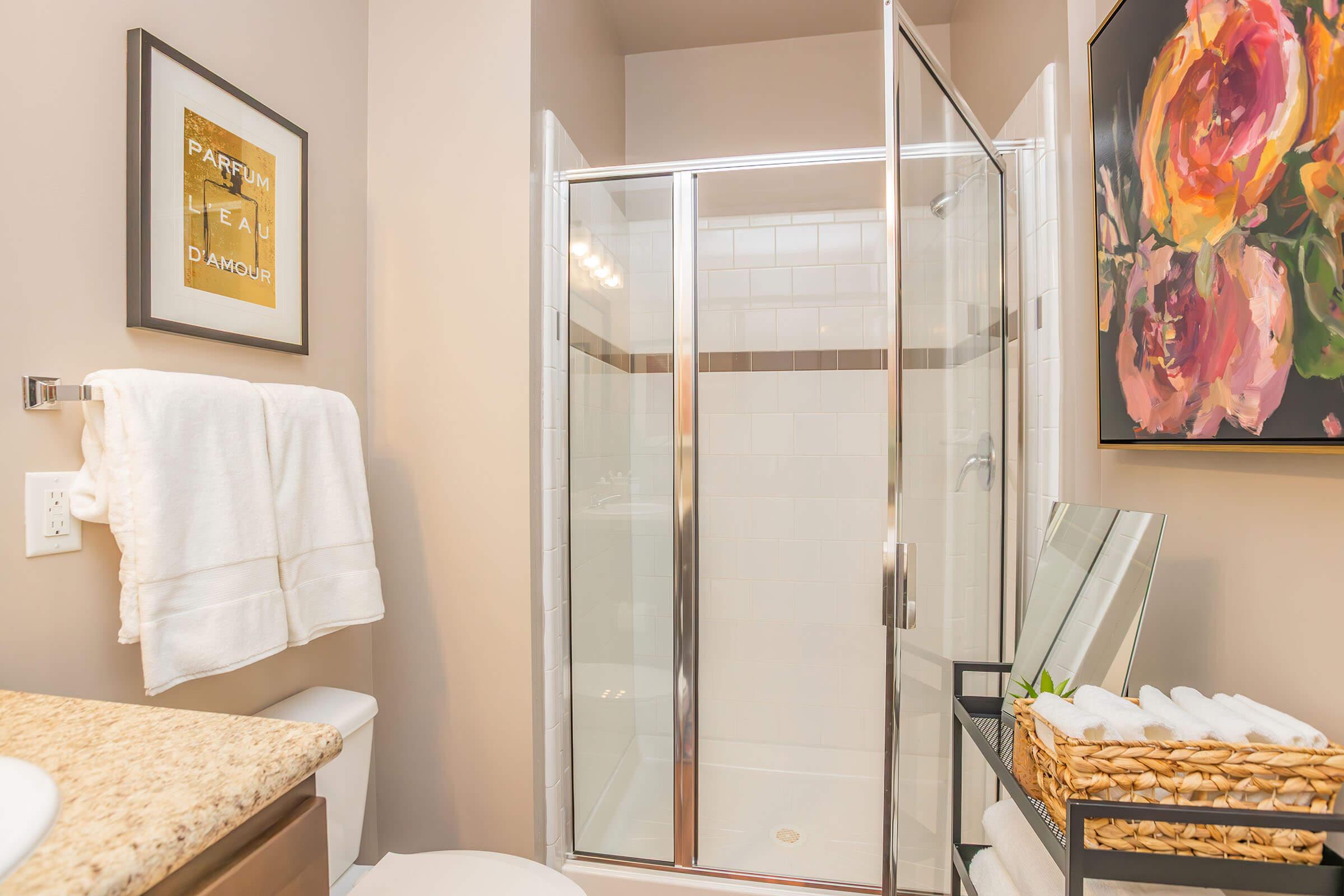 A modern bathroom featuring a glass shower enclosure, a white toilet, and a beige granite countertop. Fresh white towels hang on a towel bar, and a wicker basket contains toiletries. Decorative artwork with colorful flowers adds a touch of elegance to the wall.