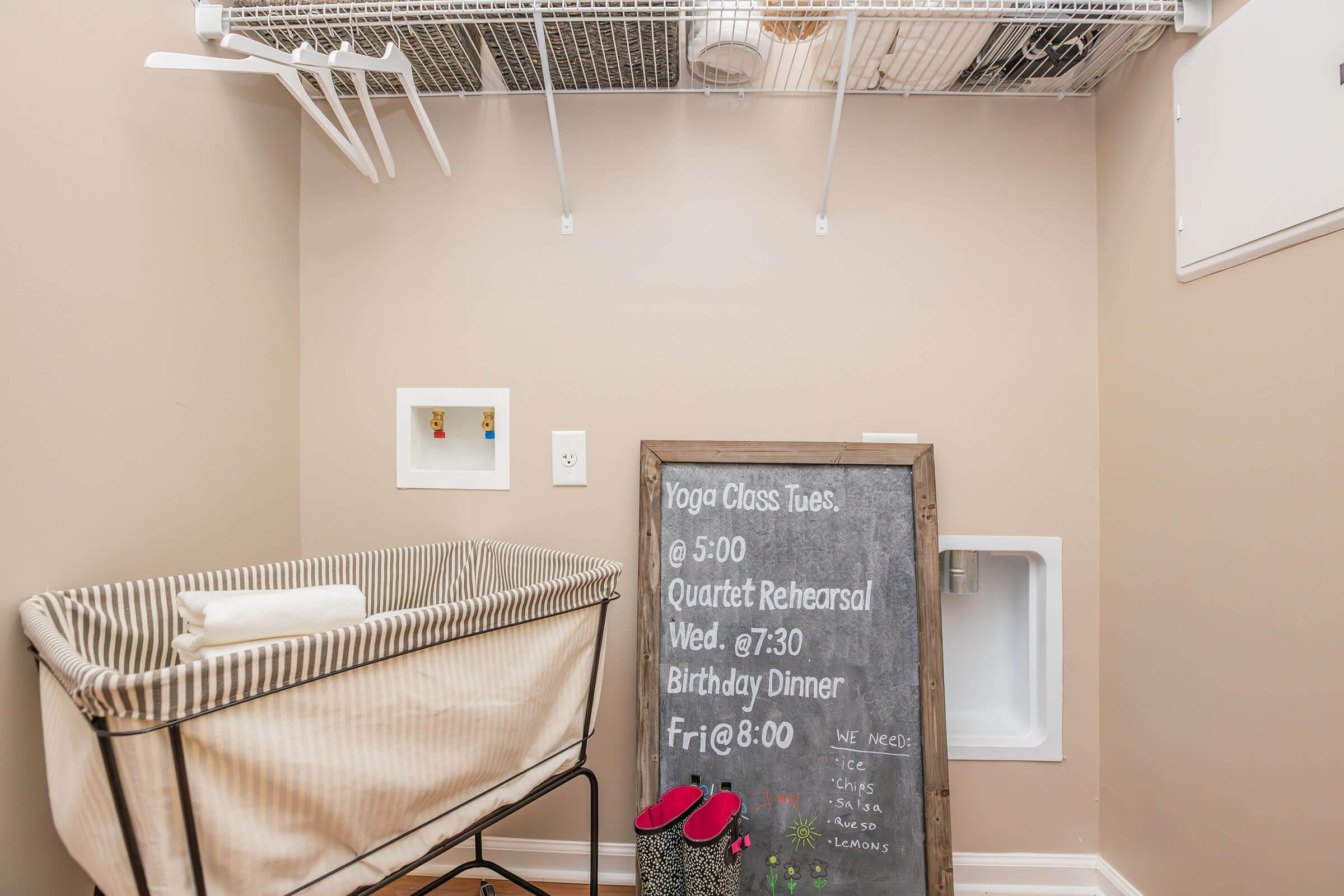A small laundry room featuring a folded baby crib on a beige wall, shelves above for storage, and a chalkboard calendar listing upcoming events: yoga class, quartet rehearsal, and a birthday dinner. The room has a clean, organized appearance with light-colored walls and a small door.