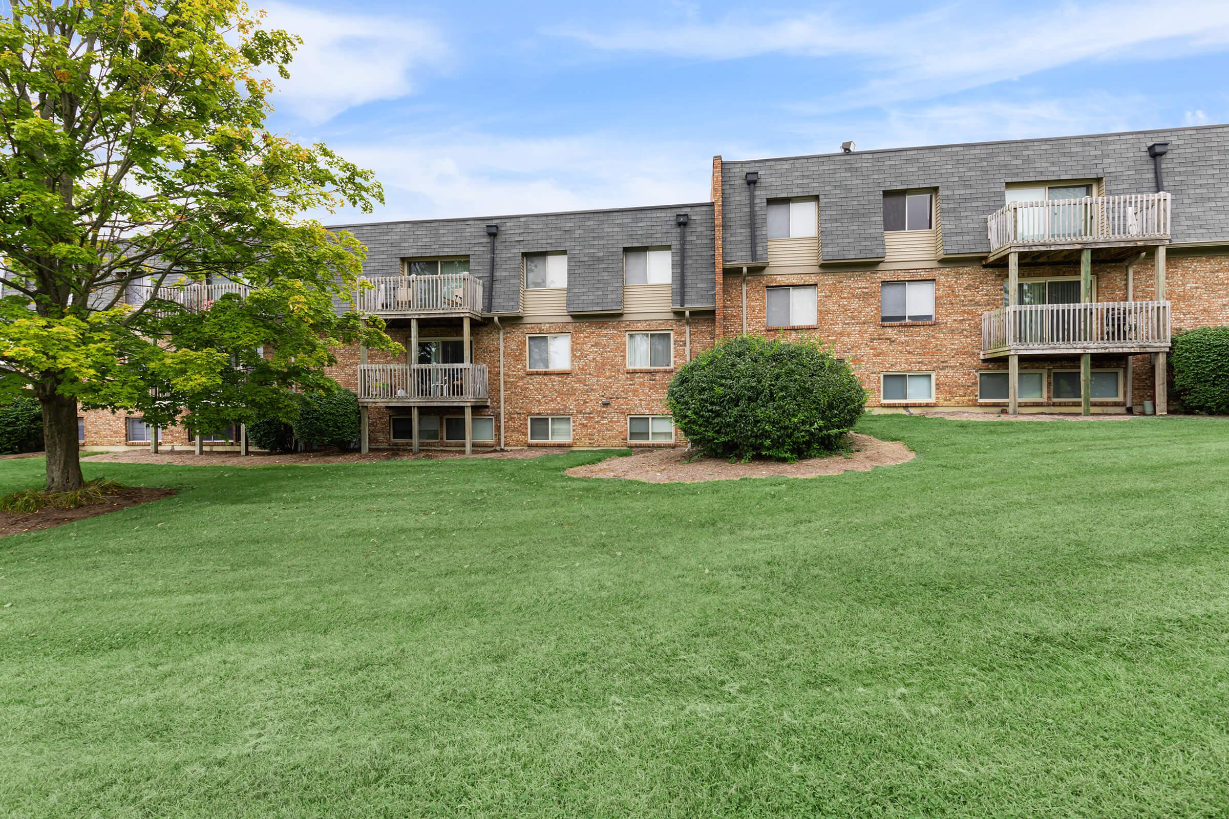 A multi-unit brick apartment building with several balconies, surrounded by a green lawn and a small tree.