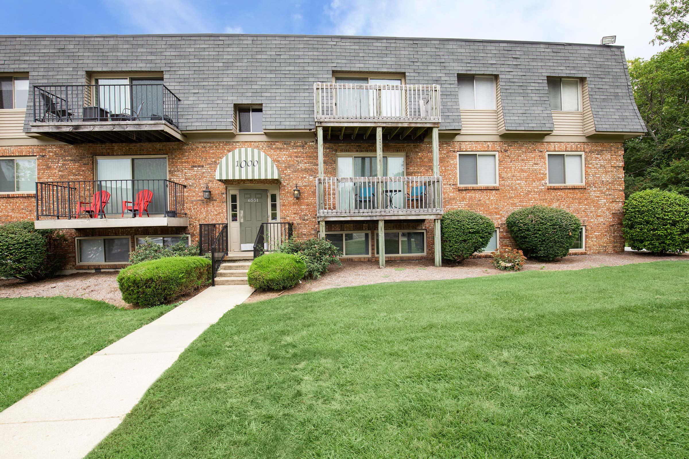 A brick apartment building with multiple balconies, surrounded by well-maintained grass and bushy landscaping. There are red chairs on the balconies, and a concrete walkway leads up to the front entrance.