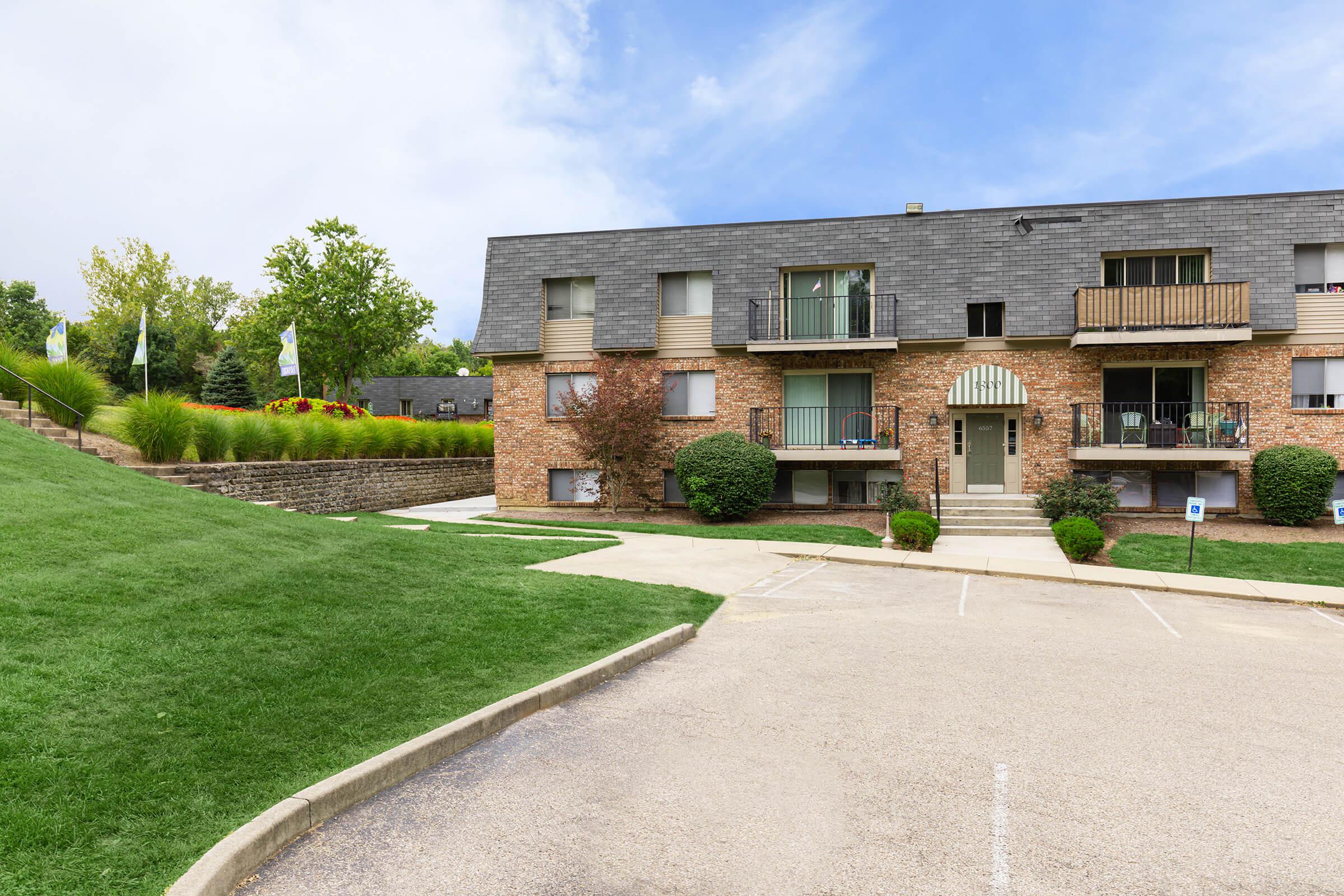 Two-story brick apartment building with a grey shingled roof, surrounded by well-maintained grass and shrubs. A parking lot is visible in front, and there is a landscaped area with decorative plants and trees nearby. Cloudy blue sky in the background.