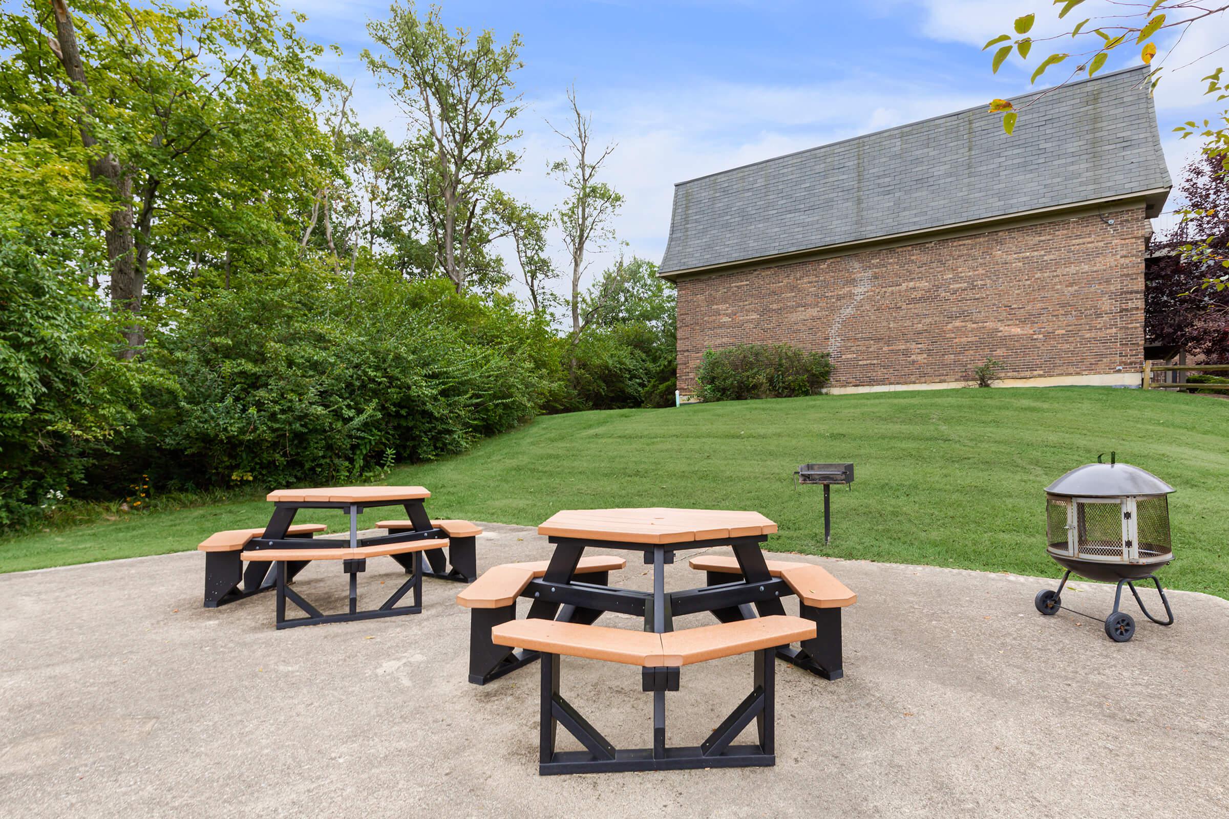 A group of wooden picnic tables on a concrete surface in a green outdoor setting, with a brick building in the background and trees surrounding the area.