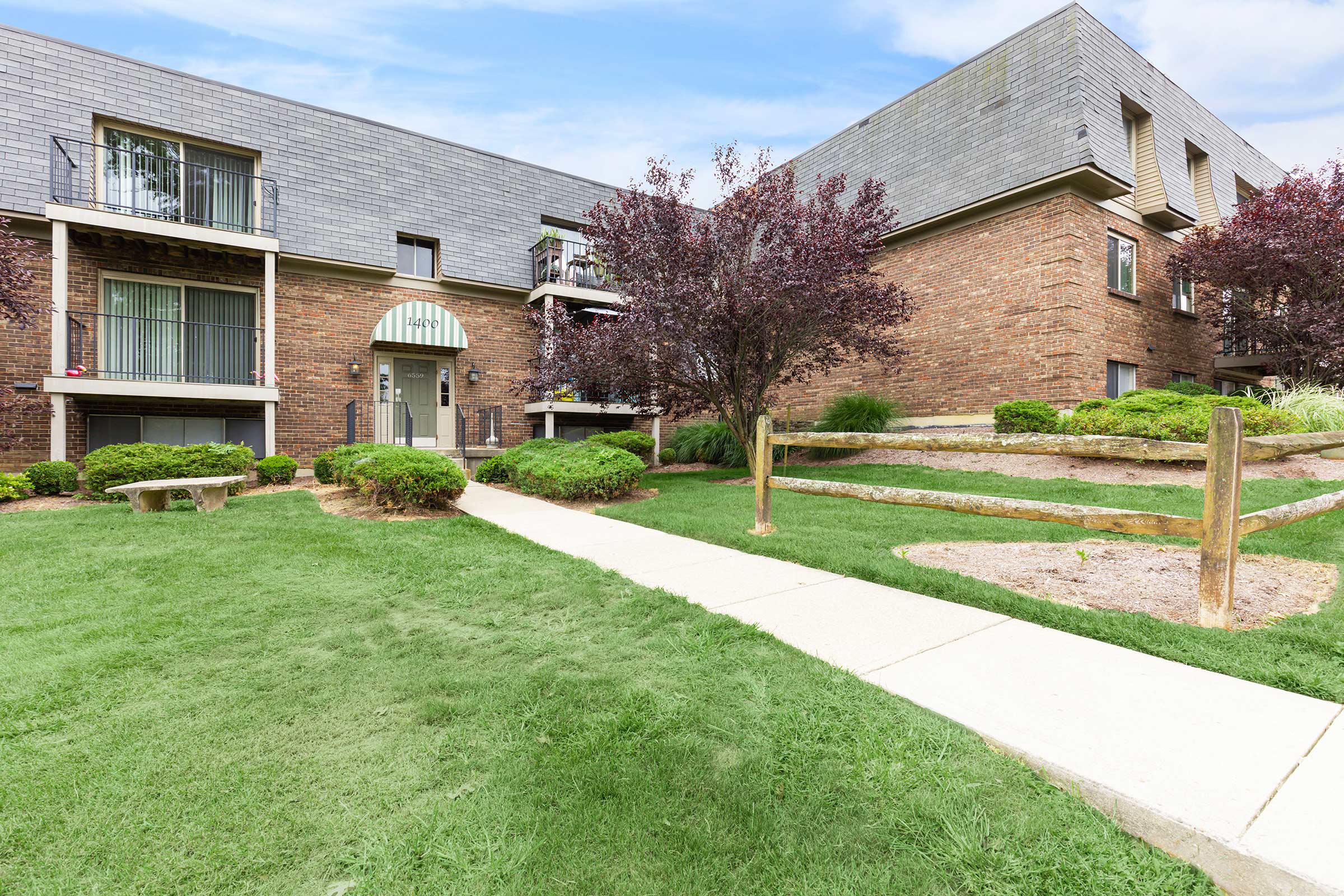 A well-maintained apartment building with brick exterior, surrounded by lush green lawns and neatly trimmed shrubs. A concrete walkway leads up to the entrance, and there are decorative trees nearby.