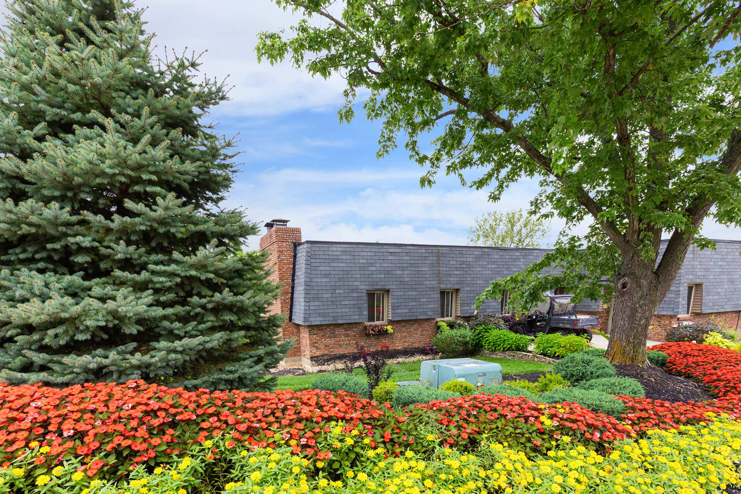 A well-maintained garden featuring vibrant red and yellow flowers in the foreground, with lush green trees and a coniferous tree to the left. In the background, there is a brick building with a sloped roof, partially obscured by the foliage and flowers, under a clear blue sky.