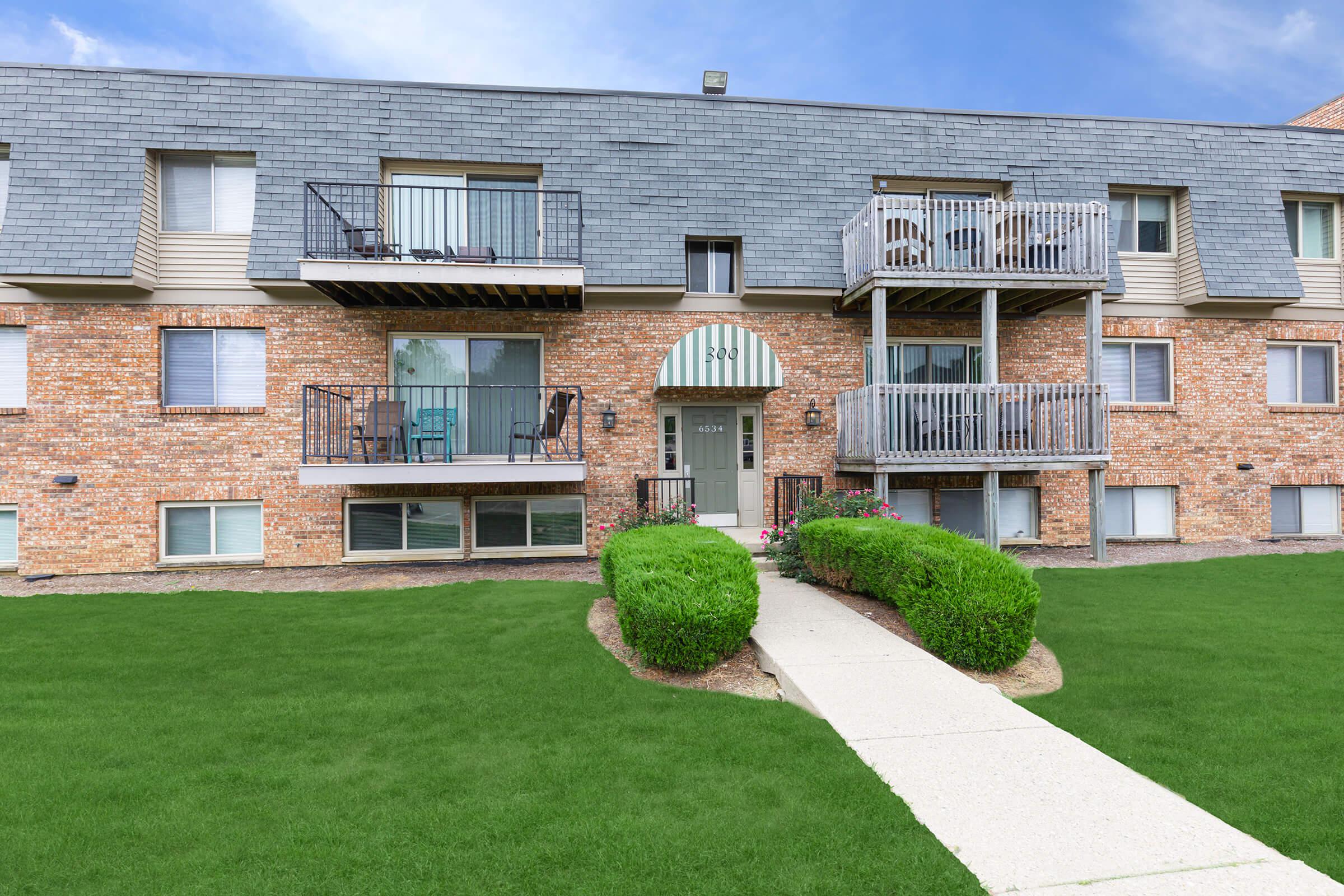 A multi-story brick apartment building with gray shingle roof and two balconies. A pathway leads to the entrance, surrounded by well-maintained green lawns and shrubs.