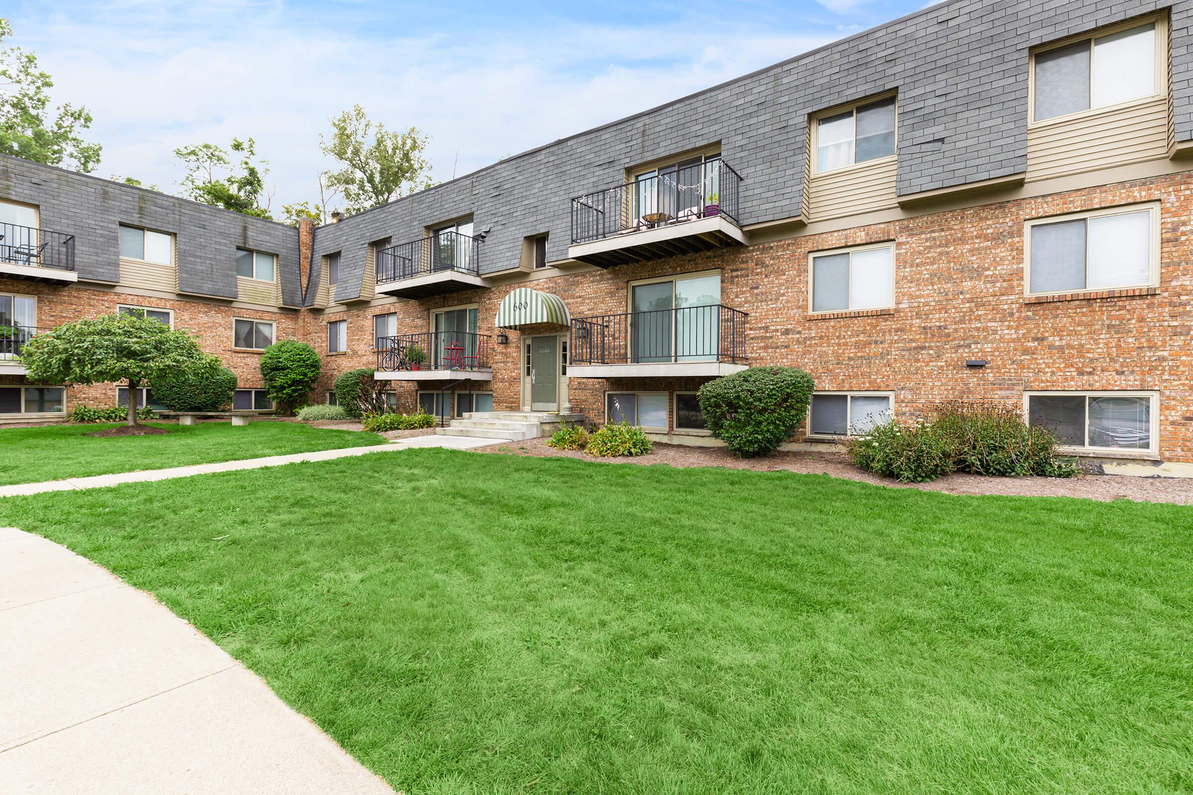 A multistory brick apartment building with balconies, surrounded by well-maintained green grass and shrubs. The building features a central entrance with an awning and large windows, and concrete pathways leading to it.