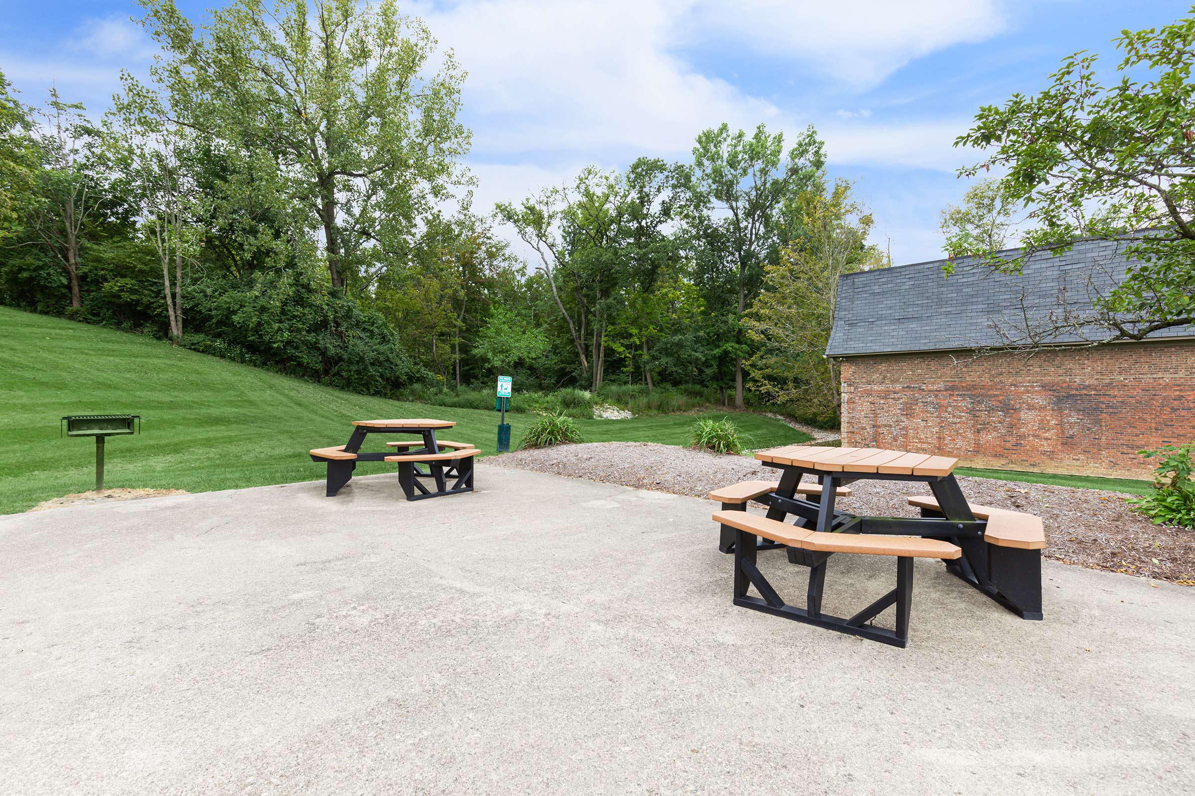 A picnic area with two wooden tables and benches set on a concrete surface, surrounded by green grass and trees in the background, next to a brick building.