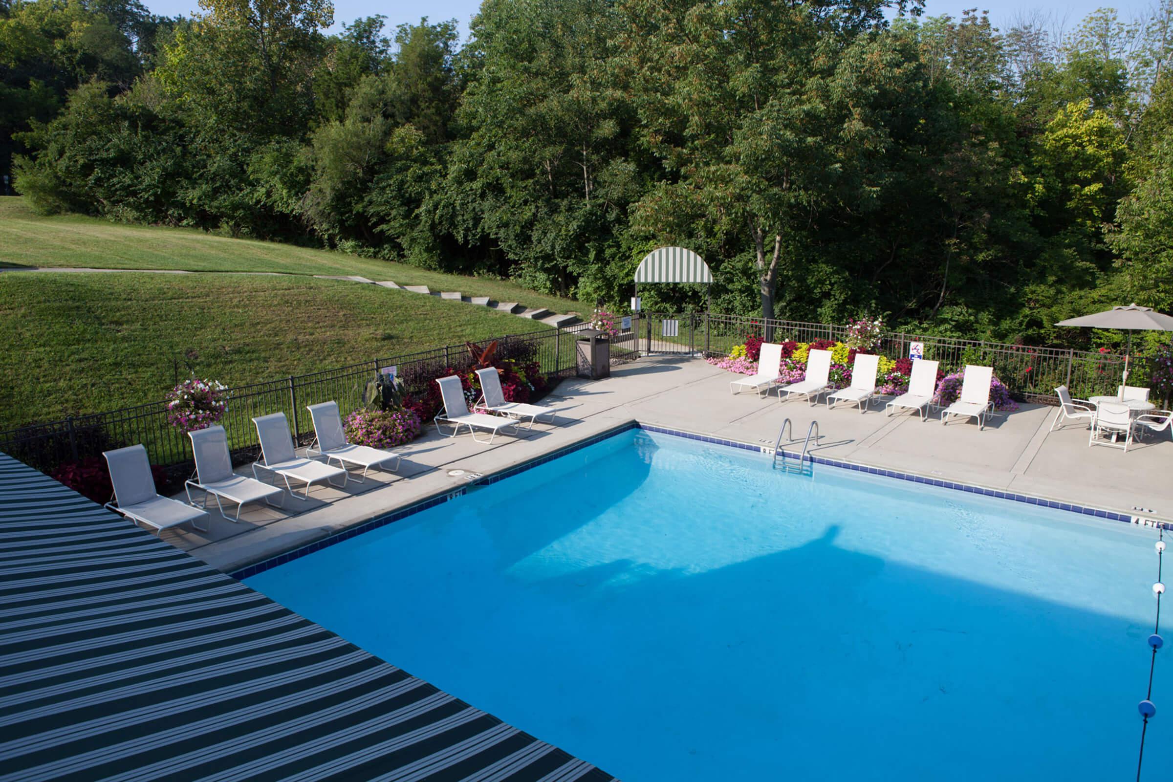 Aerial view of a swimming pool surrounded by lounge chairs and a green lawn. A shaded gazebo is visible near the pool, and there are flower planters along the edges.