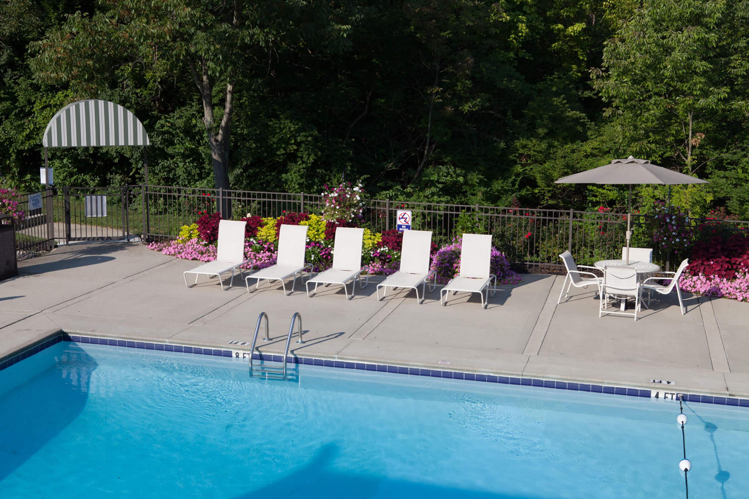 A swimming pool area featuring a blue pool, empty white lounge chairs lining the side, and a table with chairs under an umbrella. There are colorful flower beds and lush greenery in the background.