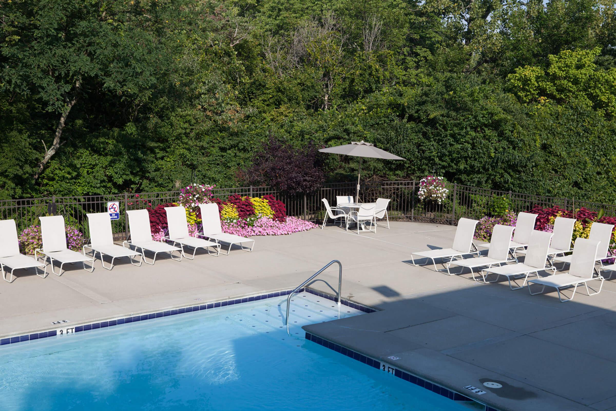 A serene outdoor swimming pool area featuring a clear blue pool, surrounded by neatly arranged white lounge chairs. Colorful flower beds fill the background, along with a shaded umbrella and additional seating. Lush greenery adds to the tranquil atmosphere.