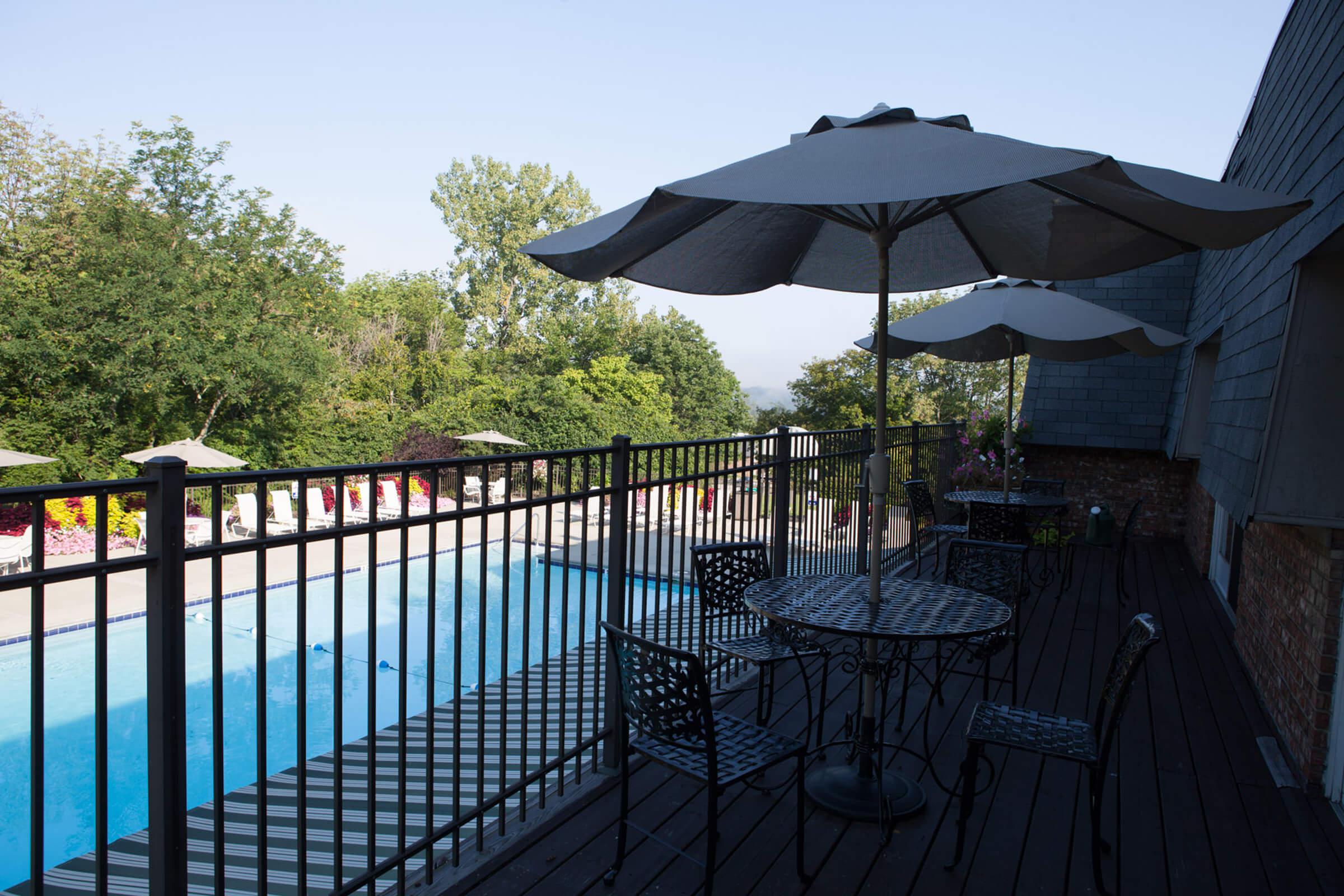 A patio area with tables and chairs under umbrellas overlooking a swimming pool, surrounded by greenery and trees.