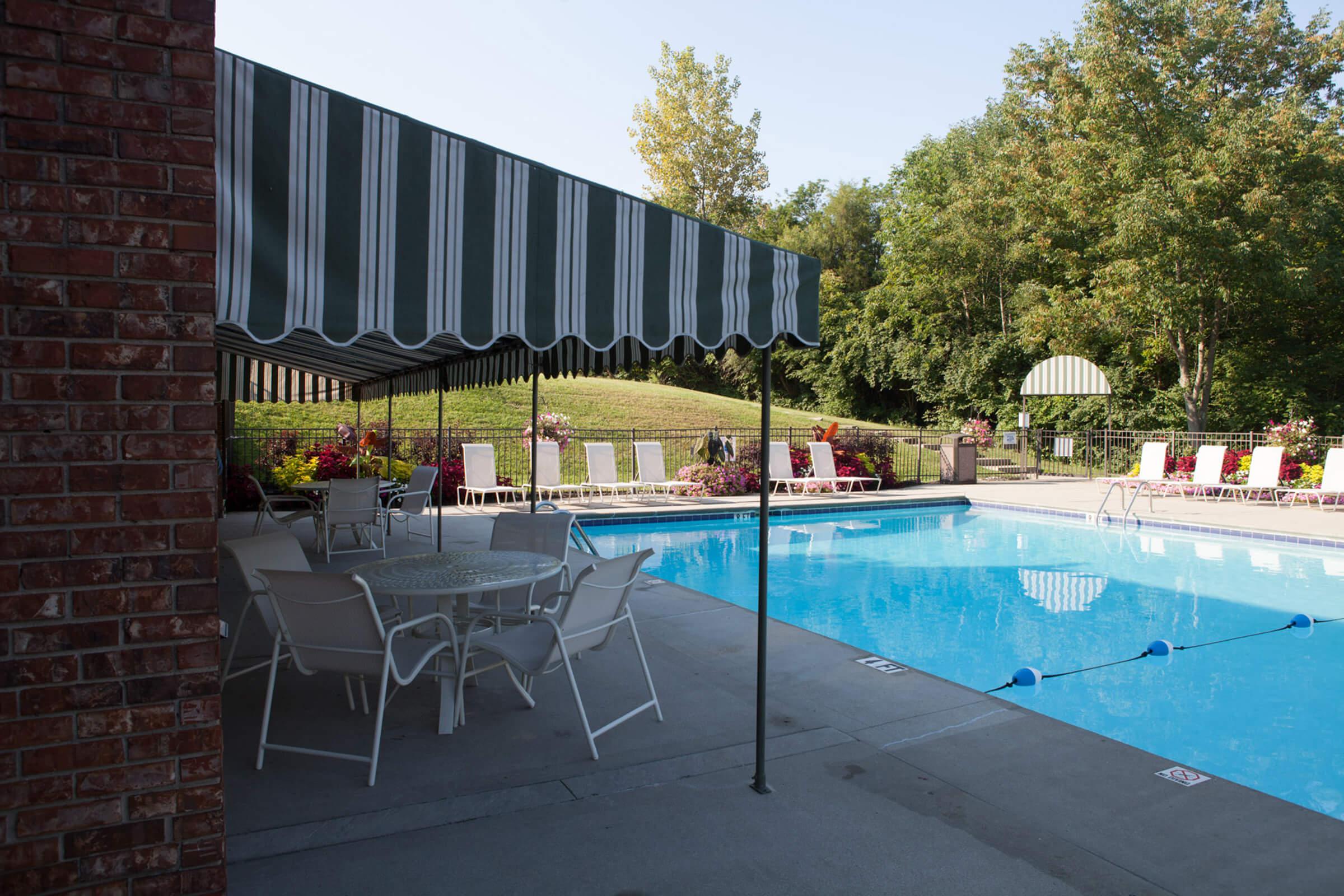 A swimming pool area featuring a clear blue pool, surrounded by lounge chairs and tables under a striped canopy. Lush green trees and grassy areas are visible in the background.