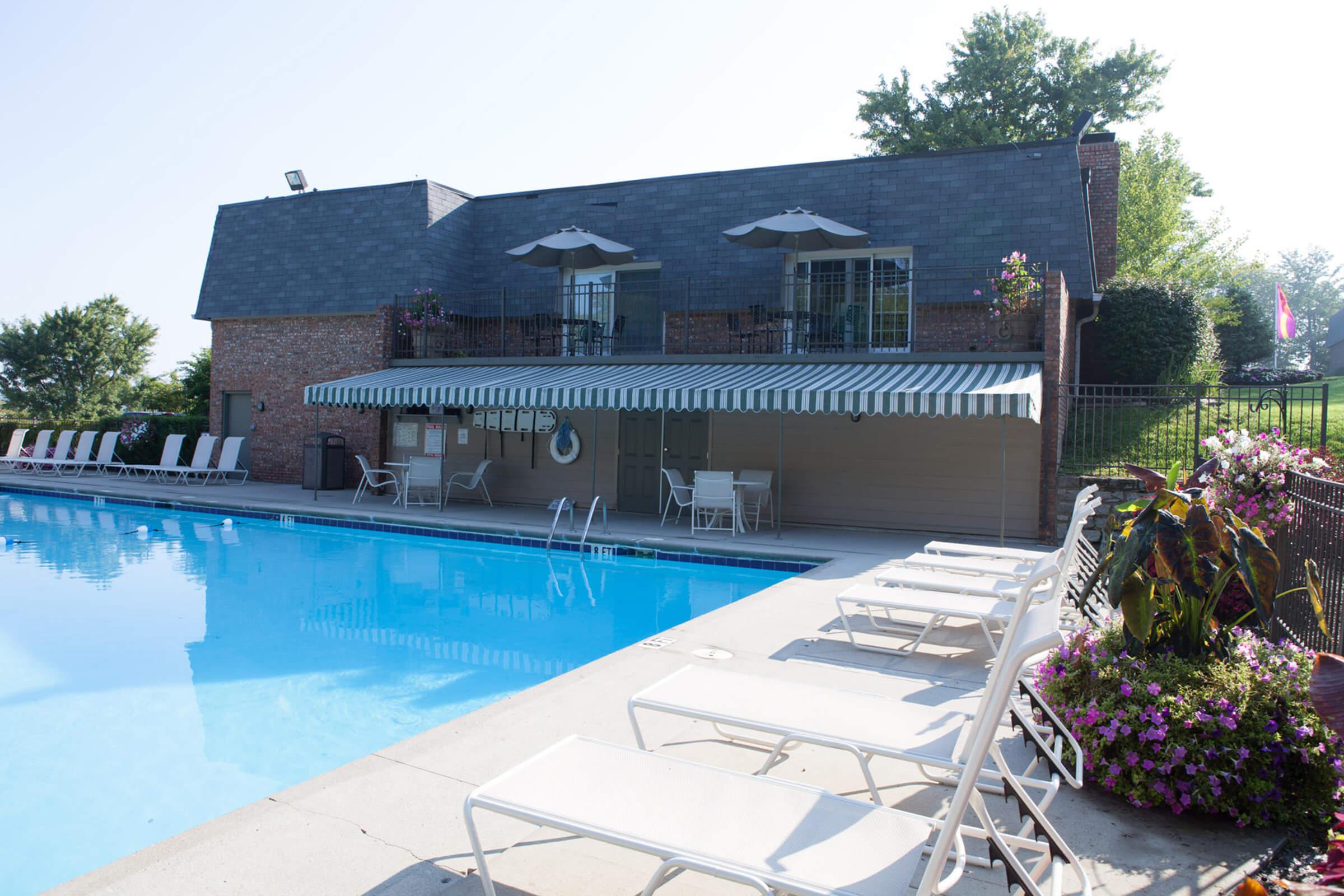 A sunny outdoor pool area featuring a clear blue pool, lounge chairs arranged neatly, and a shaded seating area with umbrellas. Surrounding the pool are blooming flowers and well-maintained greenery, along with a brick building in the background.