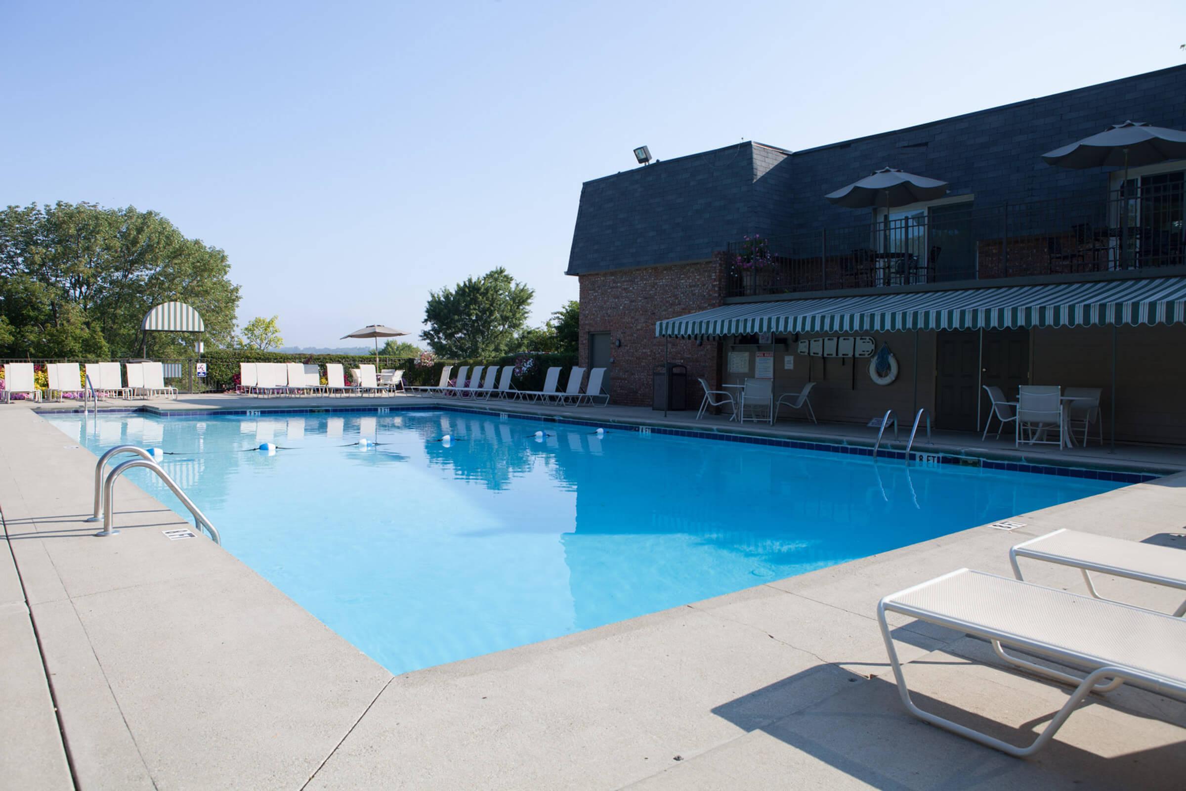 A spacious outdoor swimming pool surrounded by lounge chairs, with a shaded area and a building in the background on a clear, sunny day.