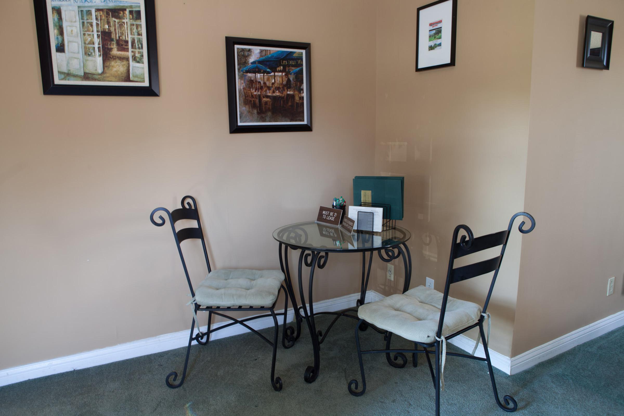 A small dining area featuring a glass-top table and two black iron chairs with cushions. The walls are adorned with framed pictures, and a decorative item sits on the table alongside a small sign. The carpeted floor is green, creating a cozy atmosphere.