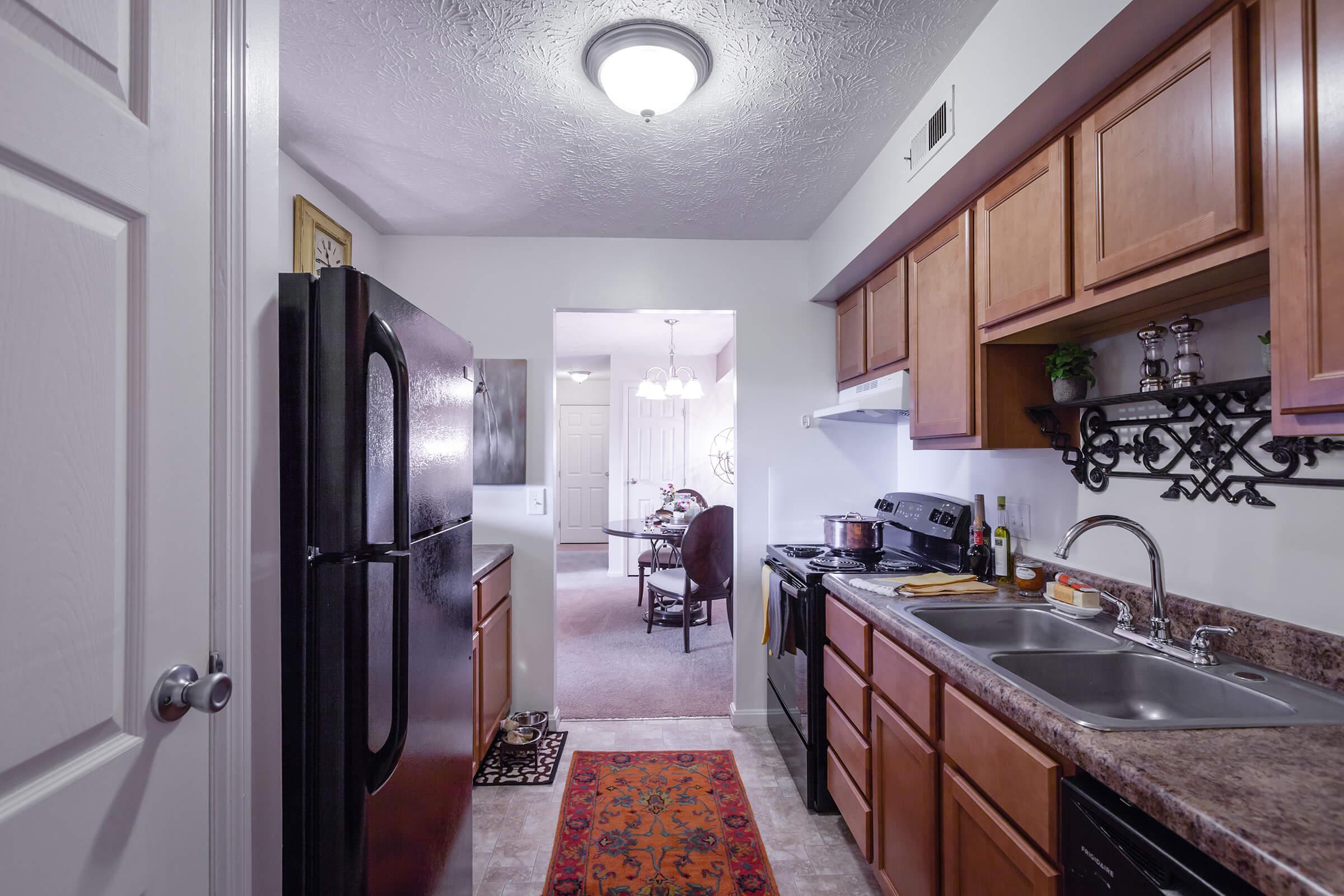 A view of a modern kitchen featuring wooden cabinets, a black refrigerator, and a double sink. Countertops are adorned with kitchen utensils, and a decorative runner is placed on the floor. The background reveals an open space leading to a dining area with a round table and chairs.