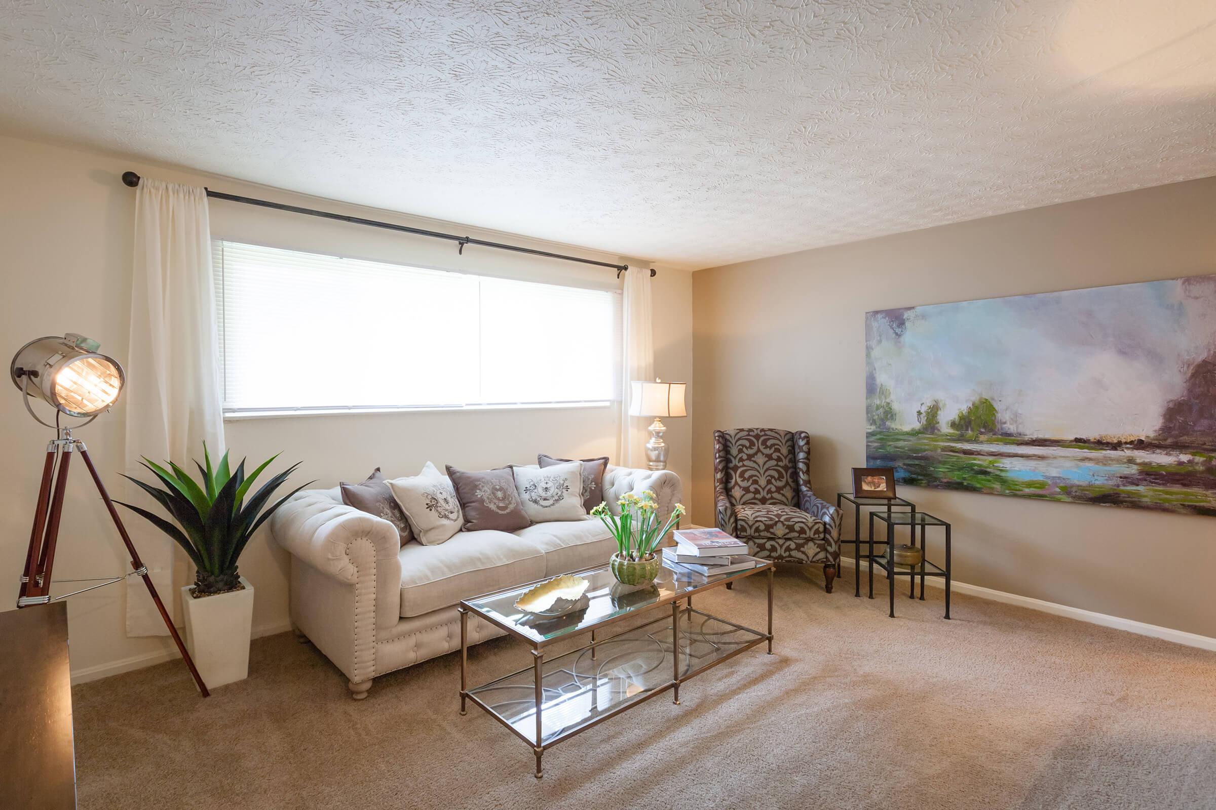 Cozy living room featuring a beige sectional sofa adorned with decorative pillows, a glass coffee table, a tall lamp, and a vibrant painting on the wall. There's a patterned chair in one corner and a potted plant on the floor, creating a comfortable and inviting atmosphere. Soft lighting and neutral tones enhance the space.