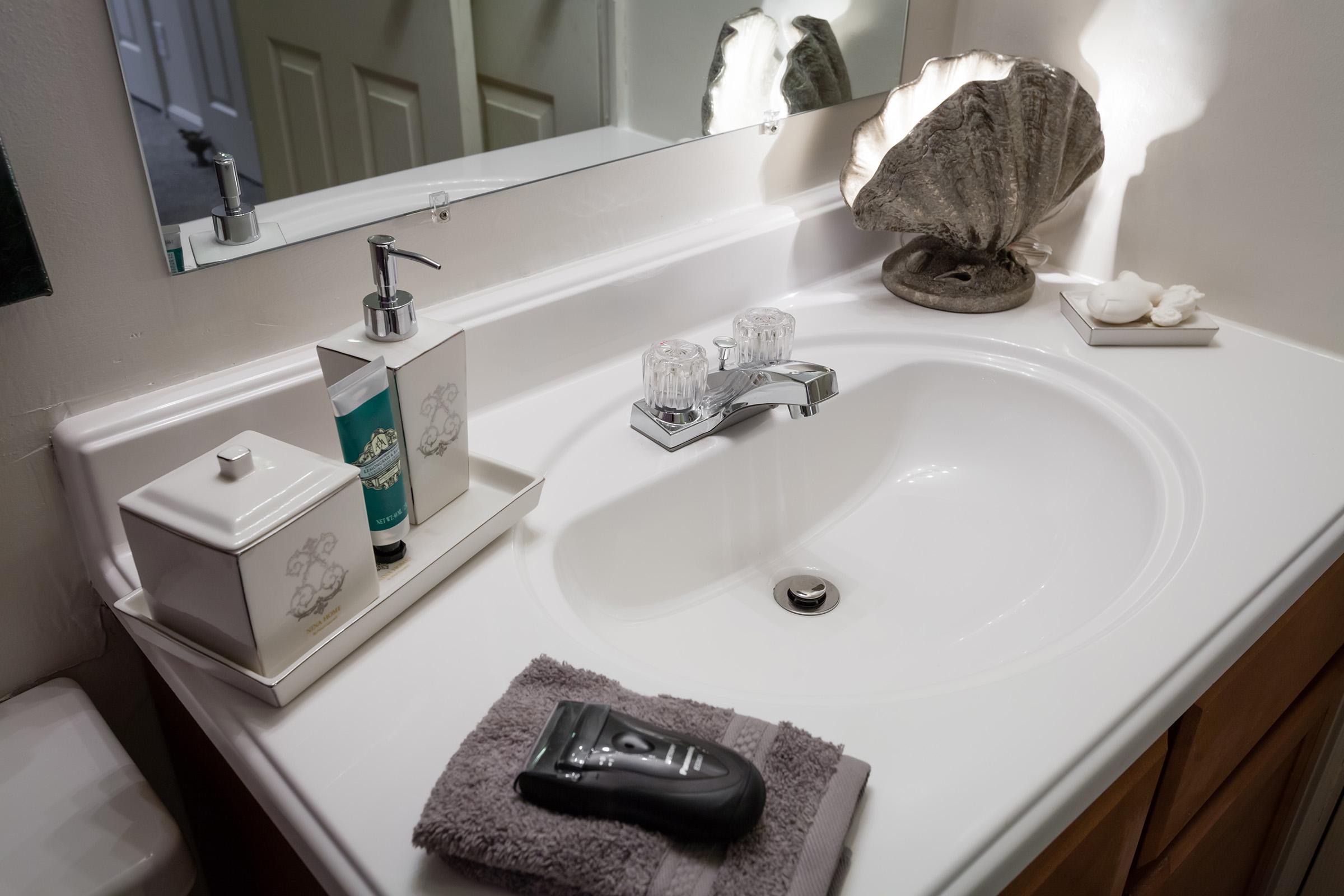 A clean bathroom sink area featuring a white basin, a decorative shell, soap dispenser, and a towel. The countertop is organized, with a toothbrush holder and a sleek shaver placed nearby, reflecting a well-maintained and tidy space. Light colors and natural elements create a serene ambiance.