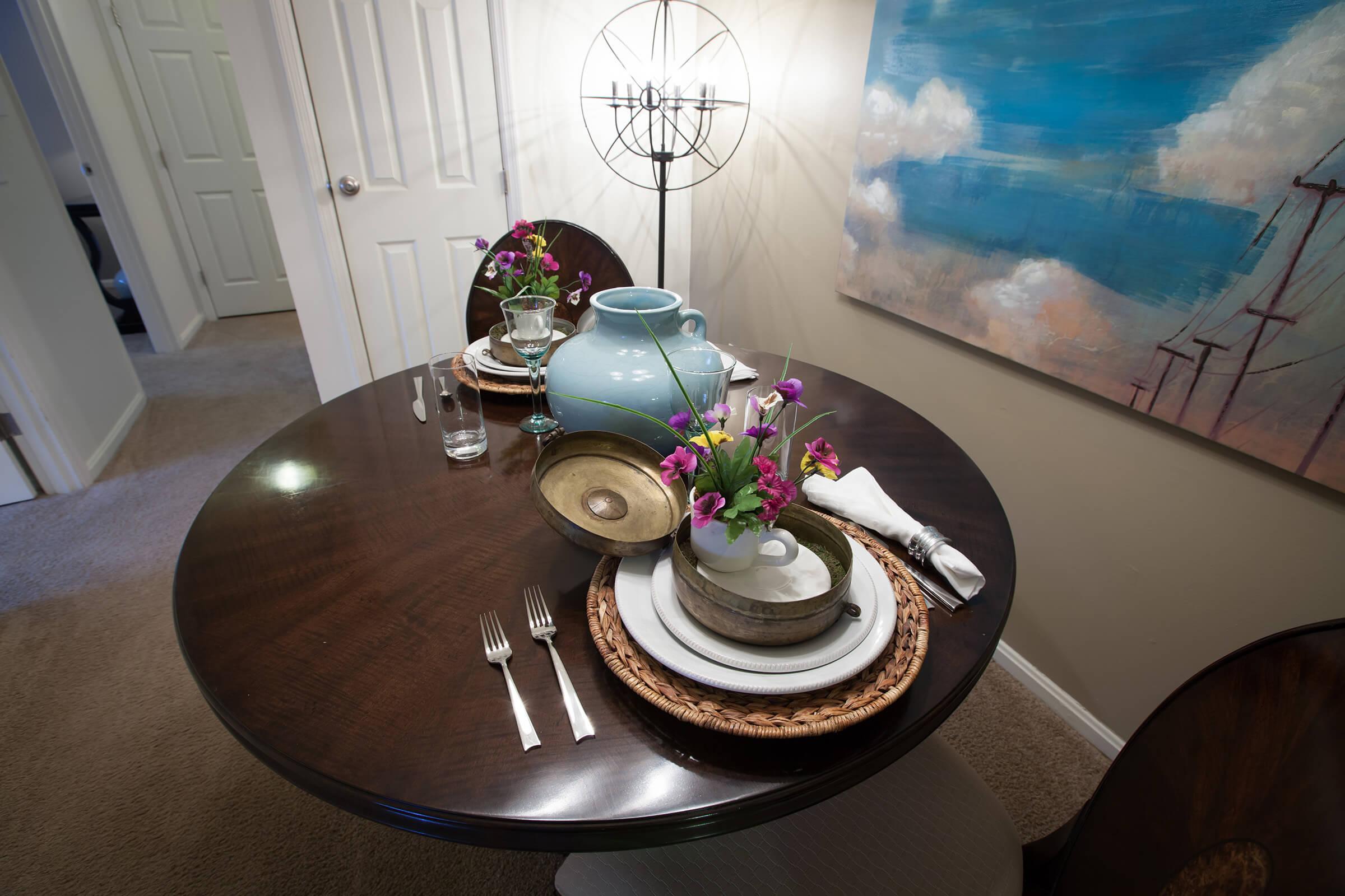 A beautifully set dining table featuring a round dark wood table with woven placemats, white dinnerware, silver cutlery, and small floral arrangements. A decorative turquoise vase adds color. A stylish lamp is illuminated nearby, and a colorful abstract painting is visible on the wall. Soft carpet underfoot.