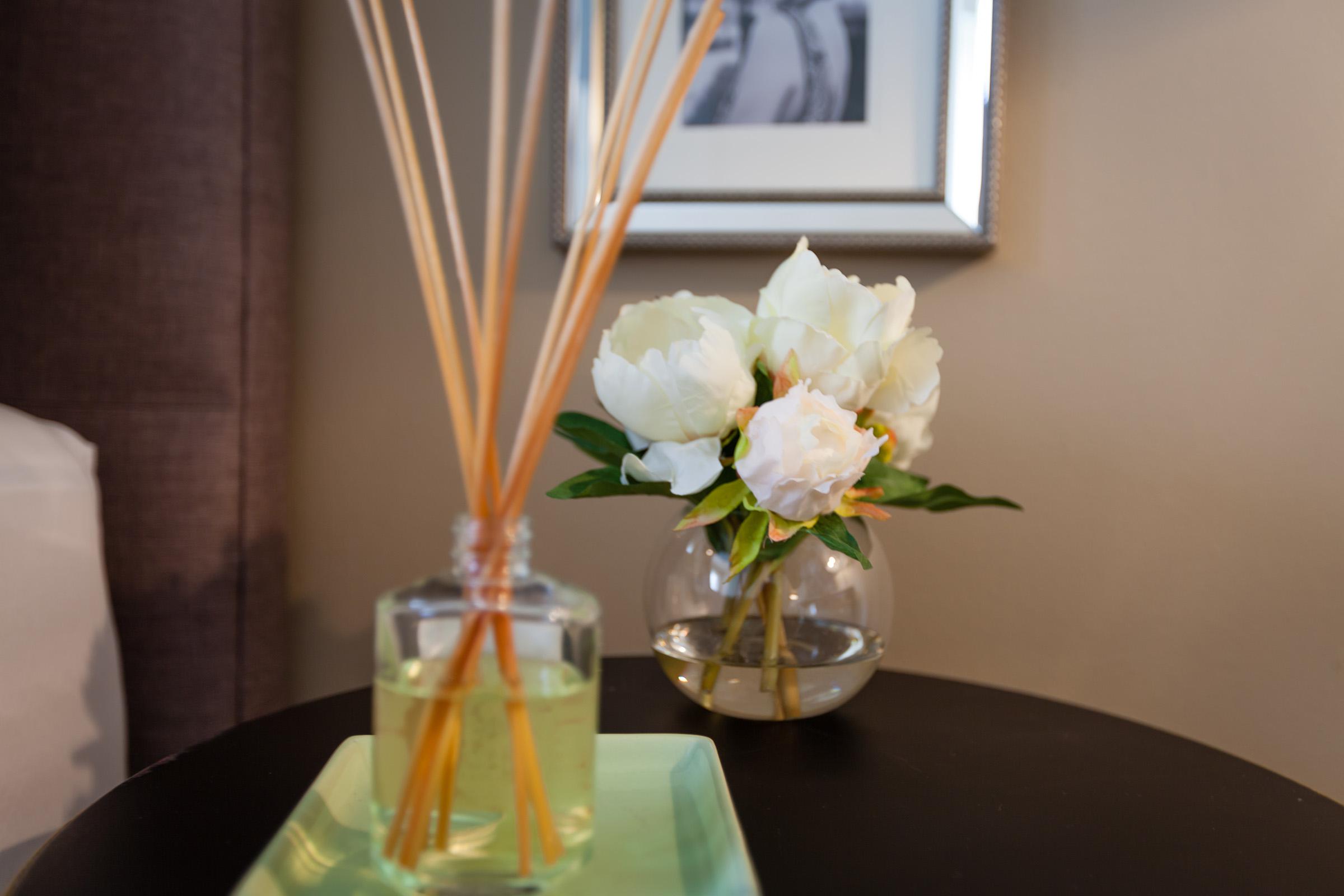 A close-up of a decorative arrangement featuring a glass vase with white peonies and green leaves, placed on a dark table. Next to the vase is a glass diffuser with wooden reeds, and a sleek green tray beneath it. The background features a subtle wall with a framed picture.