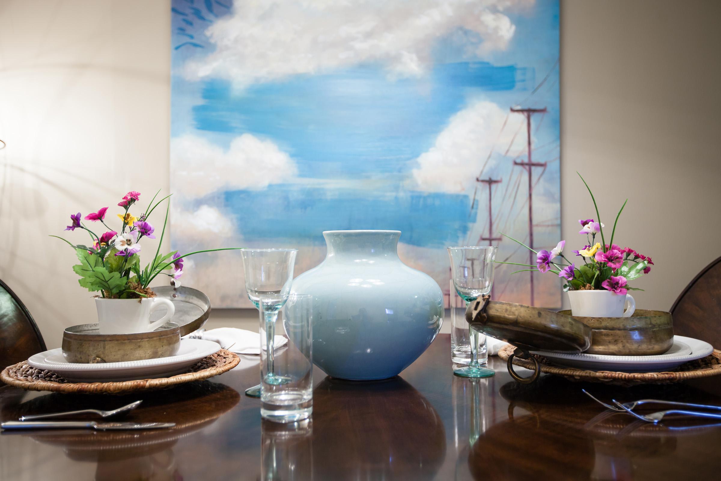 A dining table set for two, featuring elegant dishware, clear glassware, and decorative flowers in small pots. In the center, a large light blue vase stands out against a backdrop of a painting depicting clouds and sailboats on a blue sky. The atmosphere conveys a fresh and inviting dining experience.