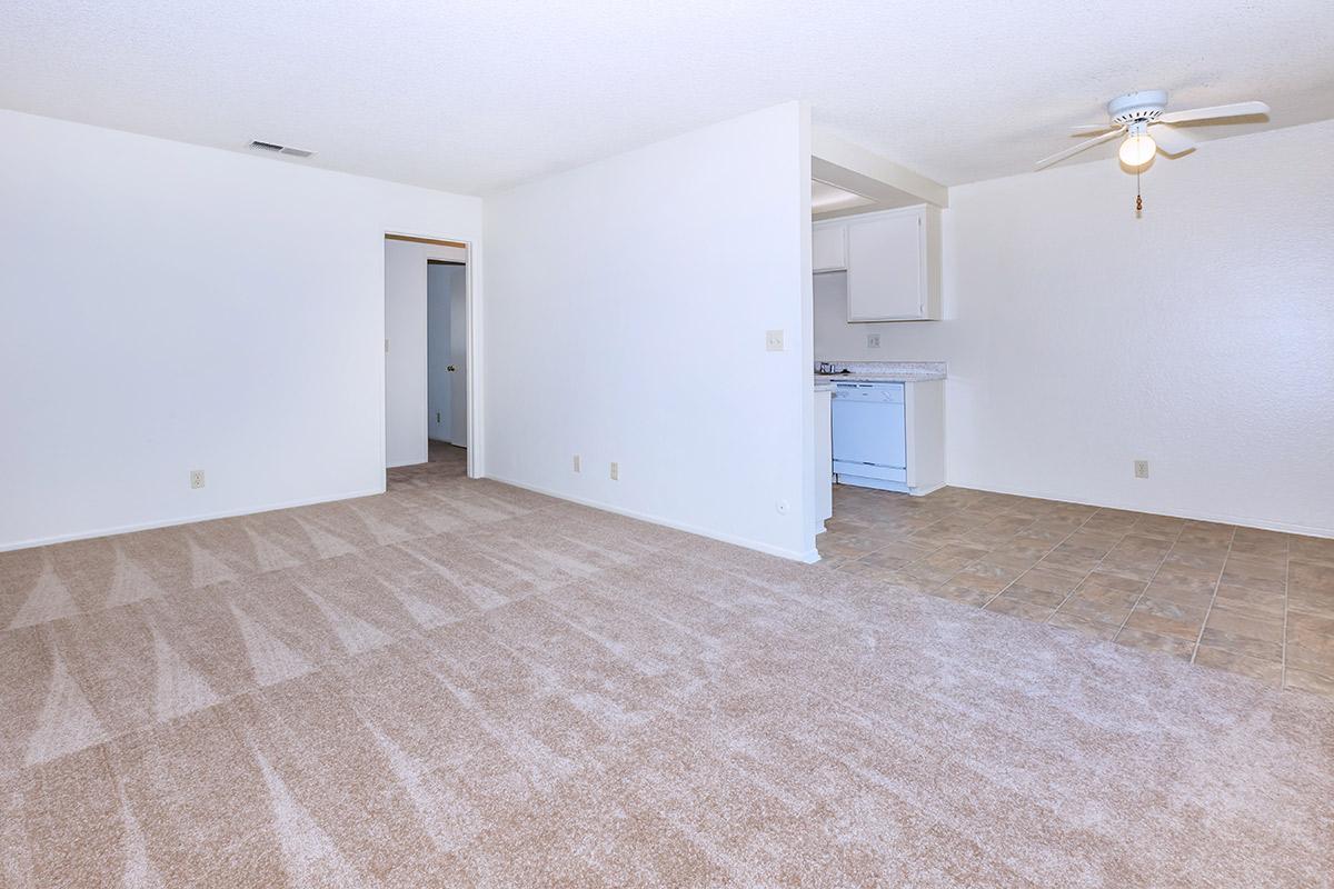 A spacious, empty living area with beige carpeting and light-colored walls. To the left, there is an entrance to another room. In the background, a kitchen area is visible, featuring light cabinetry and a tiled floor. A ceiling fan is mounted in the center of the room.
