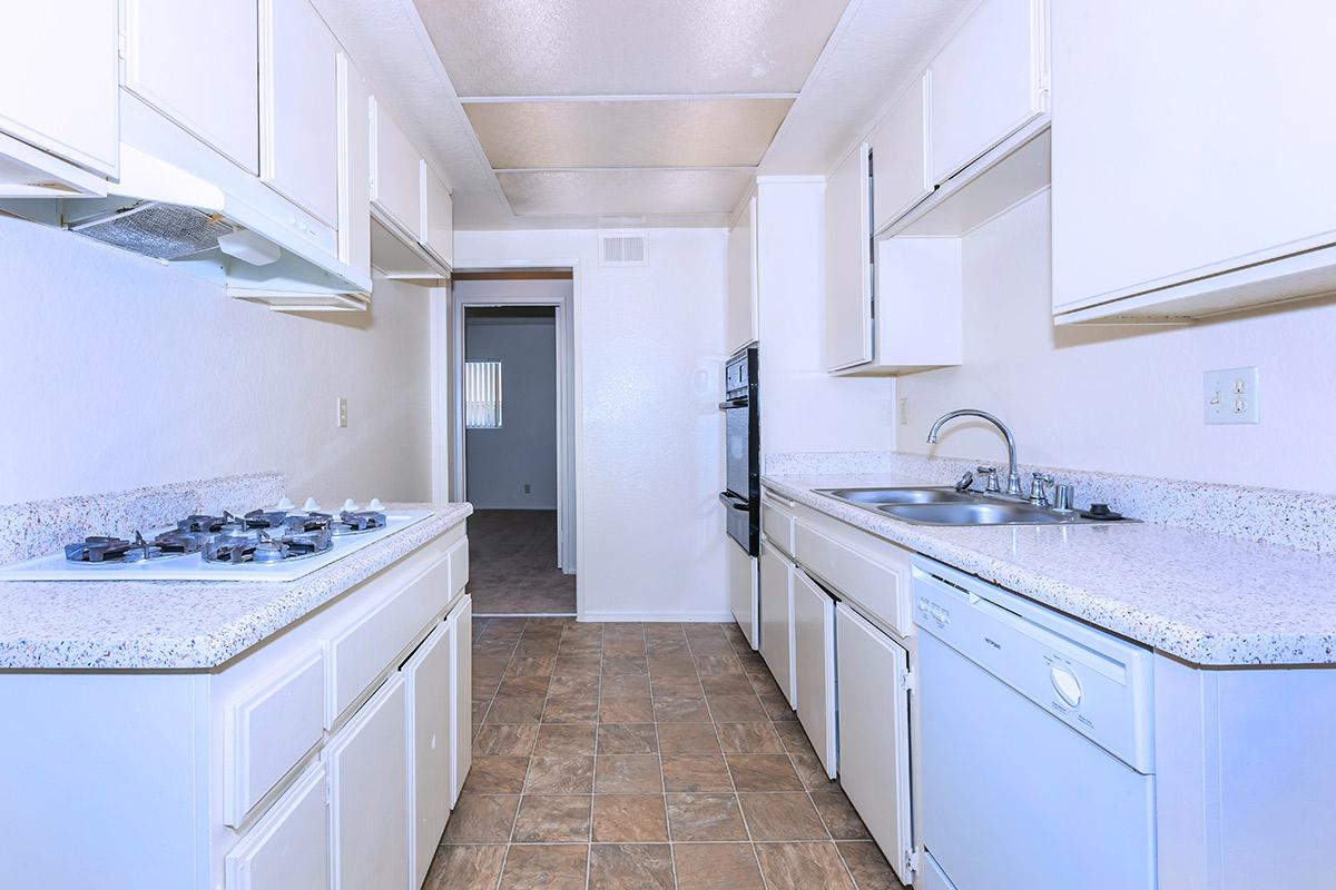 A modern kitchen featuring white cabinetry, a double sink, a gas stove with a hood, and a dishwasher. The countertop is made of light-colored granite, and the floor is tiled. The kitchen opens to a hallway leading to another room, with ample natural light and a clean, minimalist design.