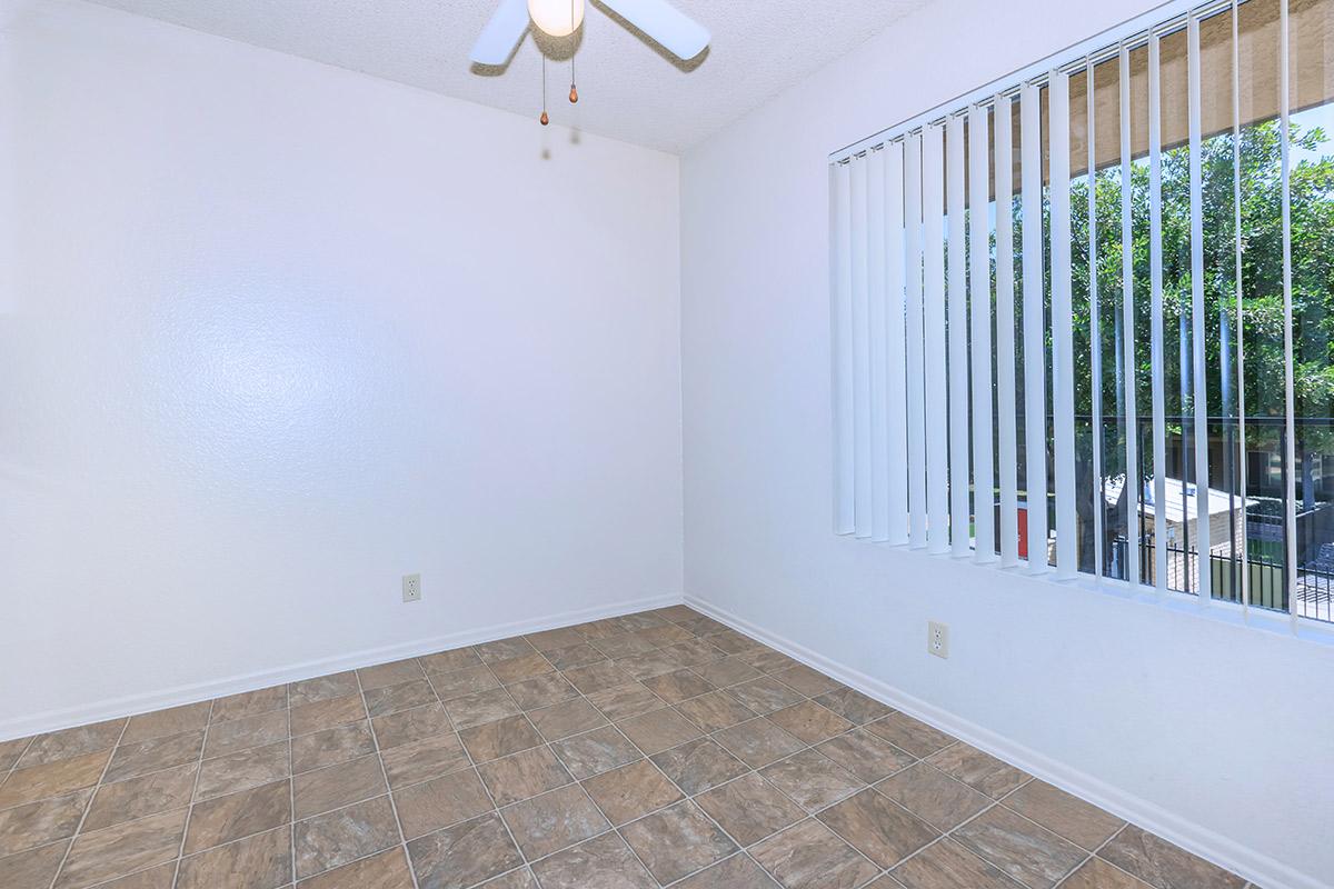 Empty room with light-colored walls and a ceiling fan. The floor is covered with tiled flooring. A window with blinds allows natural light to enter, showing greenery outside. The space appears clean and ready for furnishings.