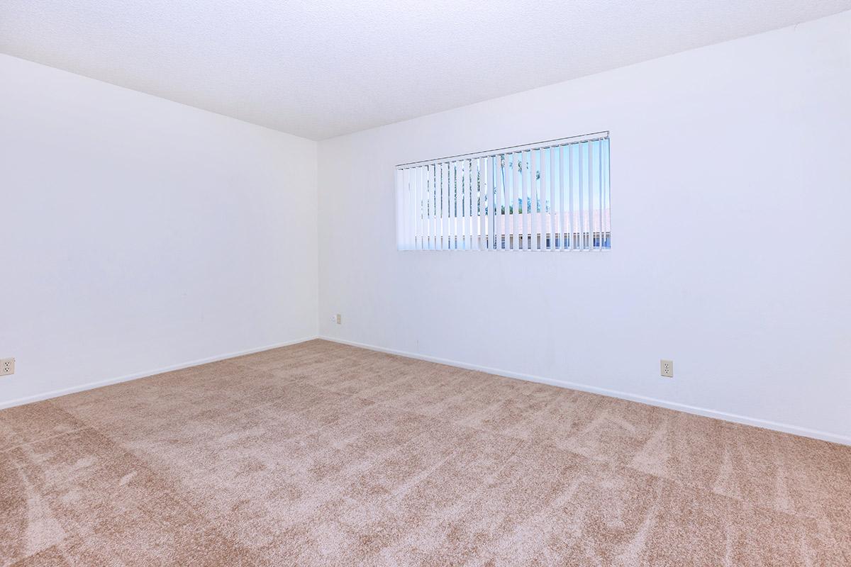 A vacant room with beige carpet and light-colored walls. There is a window with vertical blinds allowing natural light in. The room appears spacious and empty, suitable for customization or decoration.