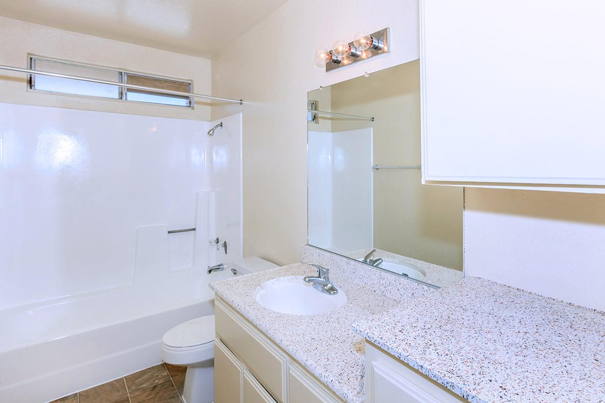 A clean, well-lit bathroom featuring a bathtub with a shower, a white toilet, and a sink with a speckled countertop. The walls are light-colored, and there's a mirror above the sink. A light fixture is mounted above the mirror, providing additional illumination.