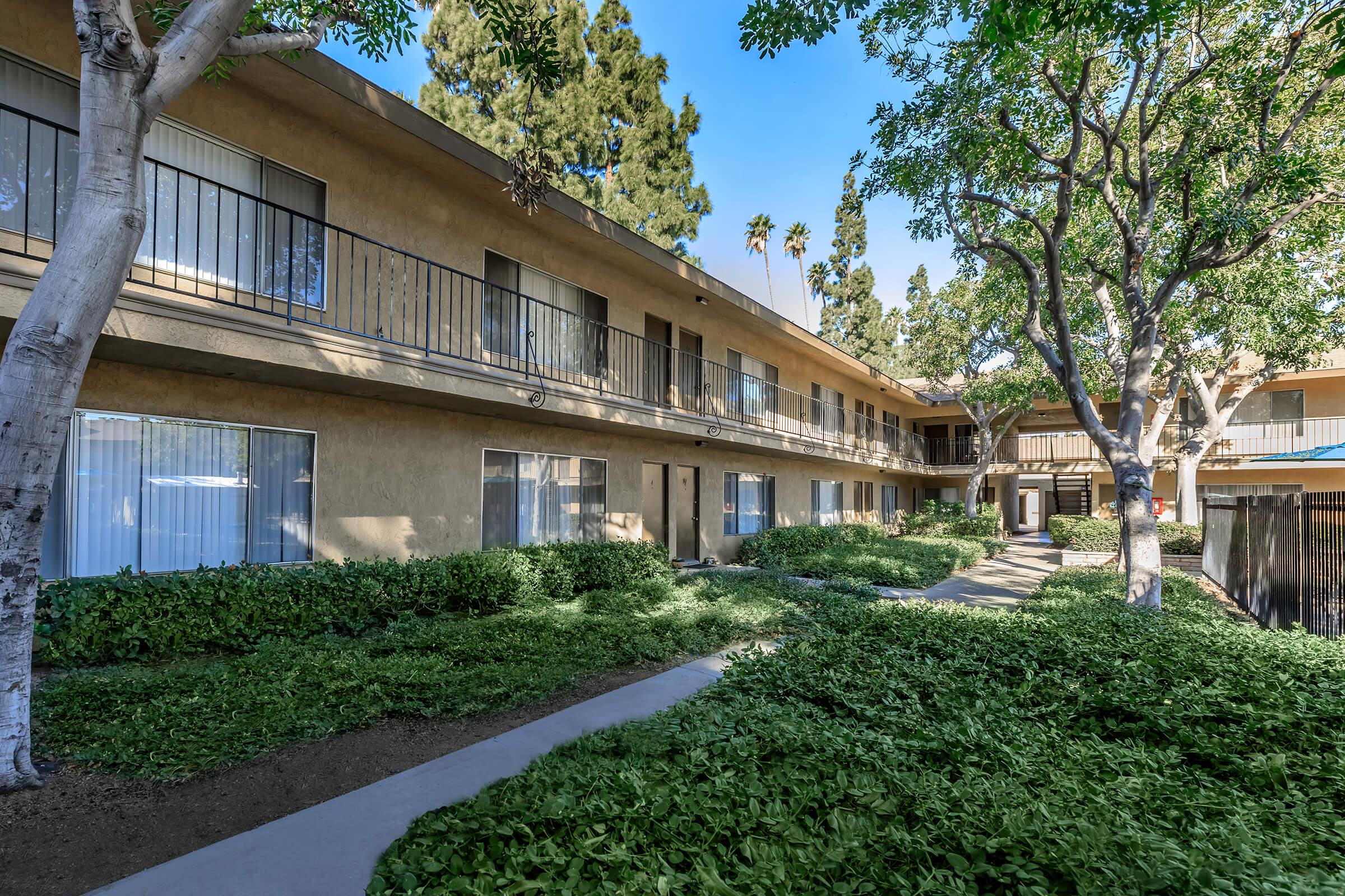 A view of a multi-story apartment complex surrounded by lush greenery. The building features multiple balconies and large windows. The pathway leads through the landscaped area, with trees providing shade and enhancing the inviting atmosphere of the property.