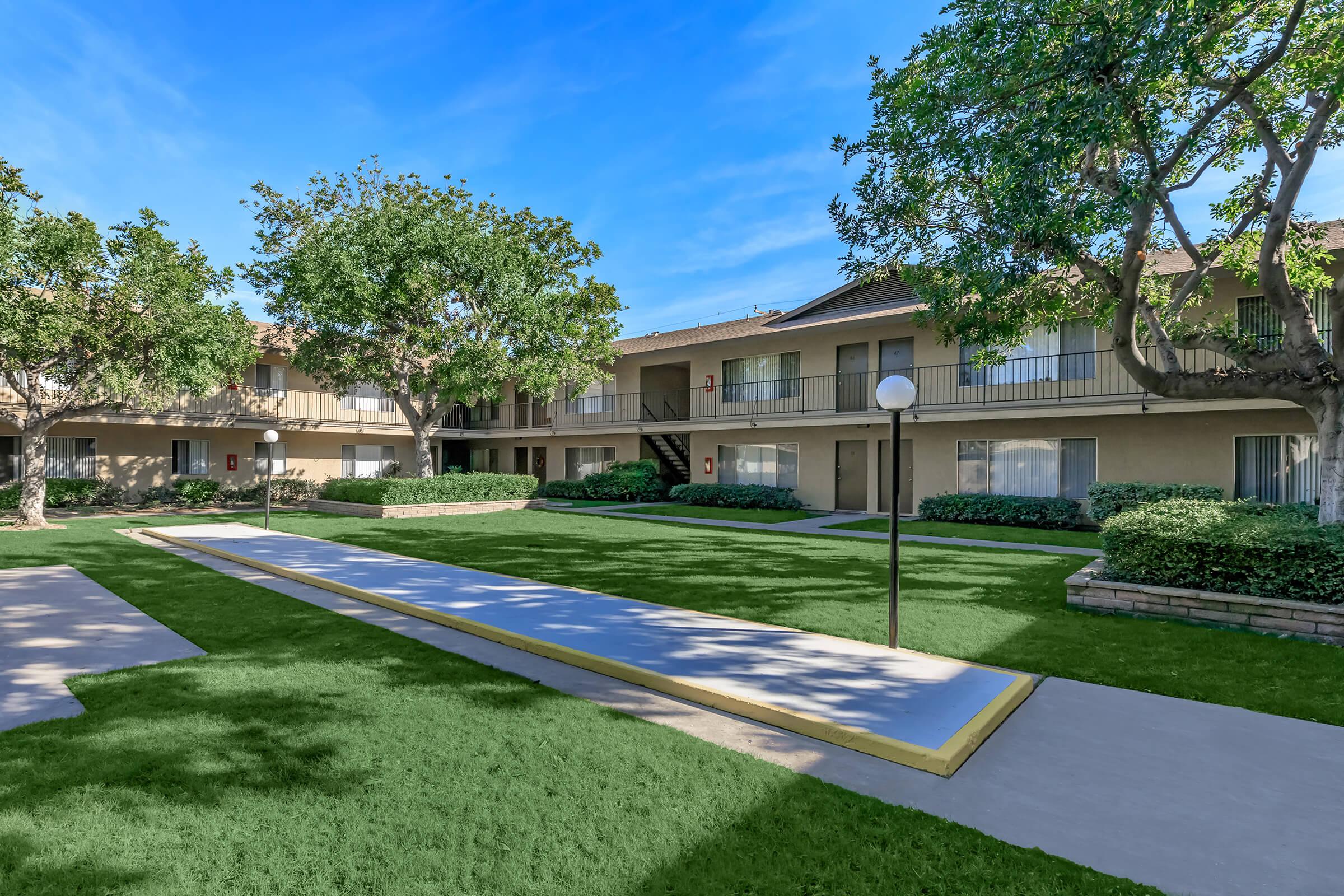 A well-maintained courtyard of an apartment complex, featuring lush green grass, several trees, and a walkway bordered by colorful bushes. The two-story buildings have balconies, and a lamp post is positioned along the path, creating a welcoming outdoor space. Bright blue sky overhead.
