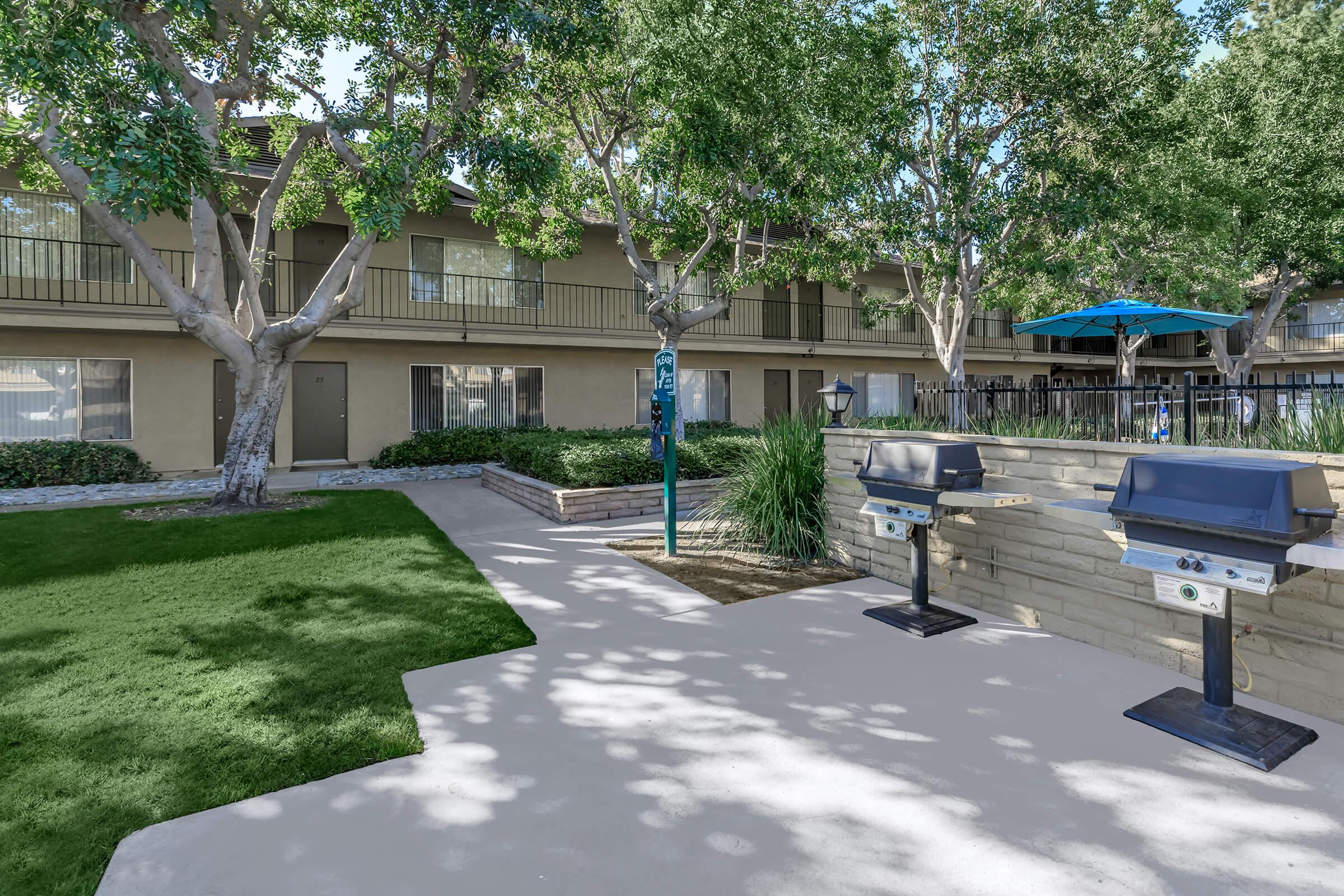 Outdoor area of an apartment complex featuring a well-kept lawn, two barbecue grills, and shaded seating arrangements under blue umbrellas. The background includes multi-story buildings with balconies, surrounded by trees, creating a welcoming and recreational atmosphere.