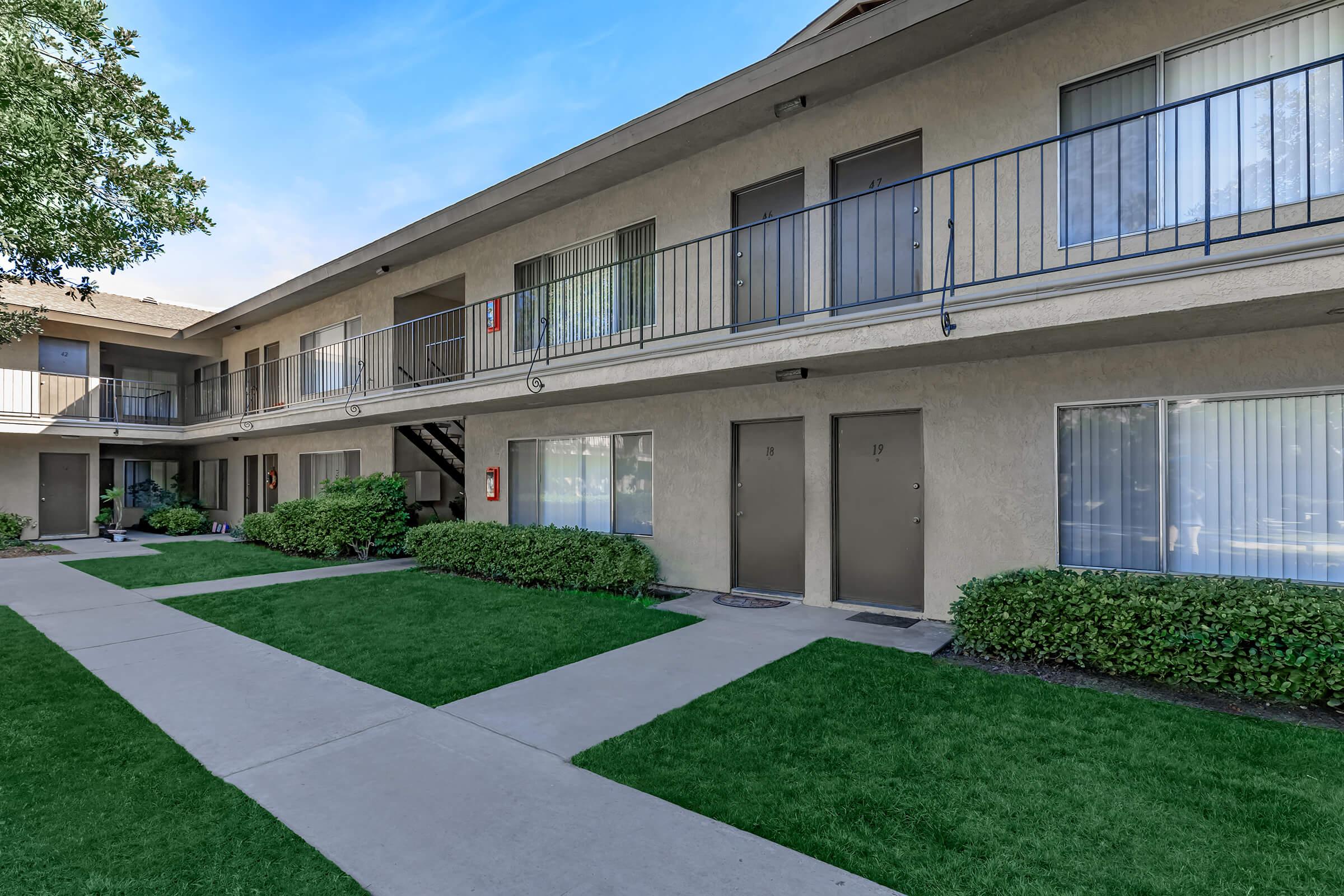 A view of a multi-unit apartment complex featuring two stories. The building has several doors with balconies, surrounded by green lawn and landscaping. A walkway leads through the grass, providing access to the entrances. There are emergency exit signs visible on the walls.