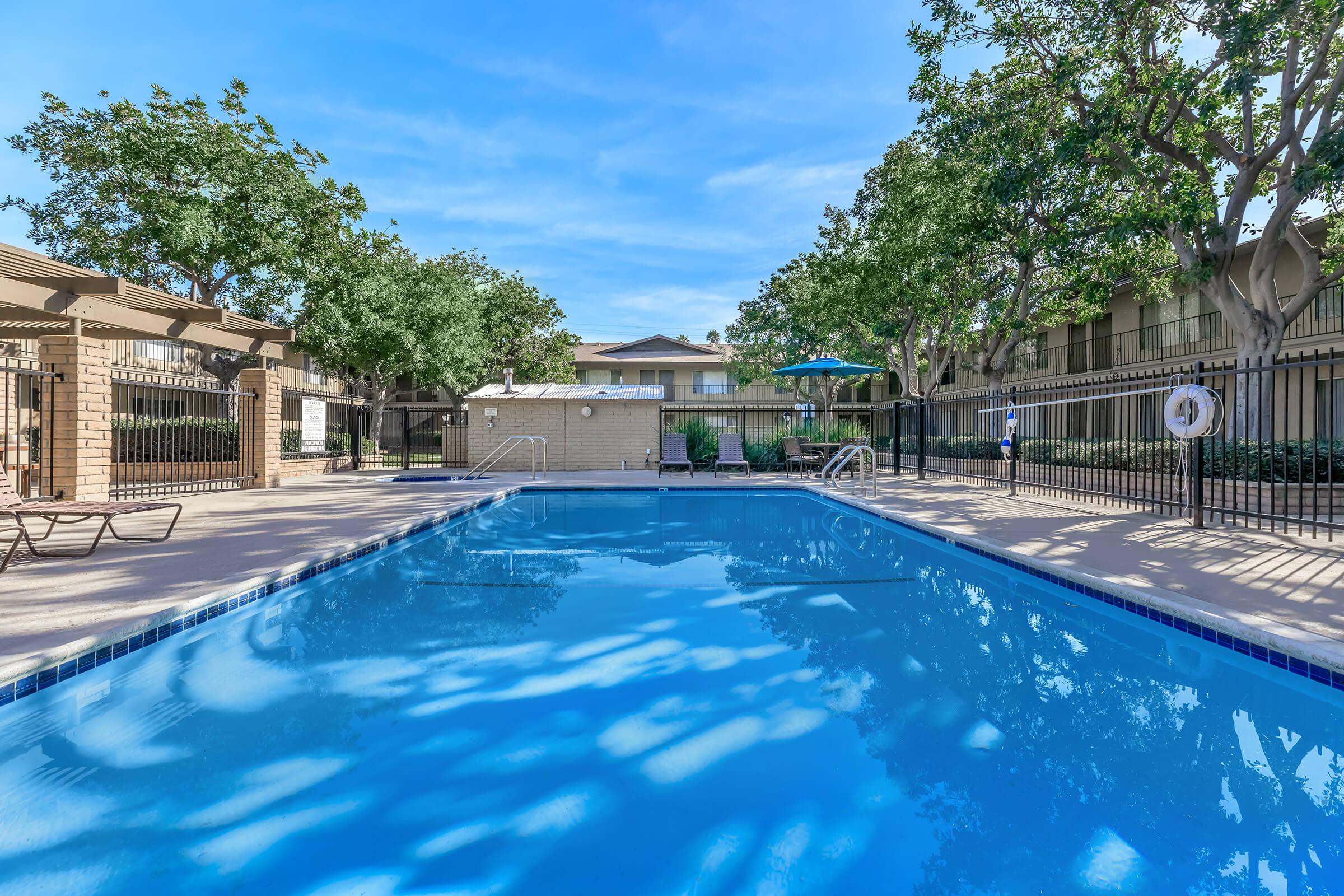 A clear blue swimming pool surrounded by lush trees and greenery, with lounge chairs and umbrellas nearby. The pool area is well-maintained, featuring a fence and a building in the background. The sky is bright and sunny, creating a relaxed, inviting atmosphere.