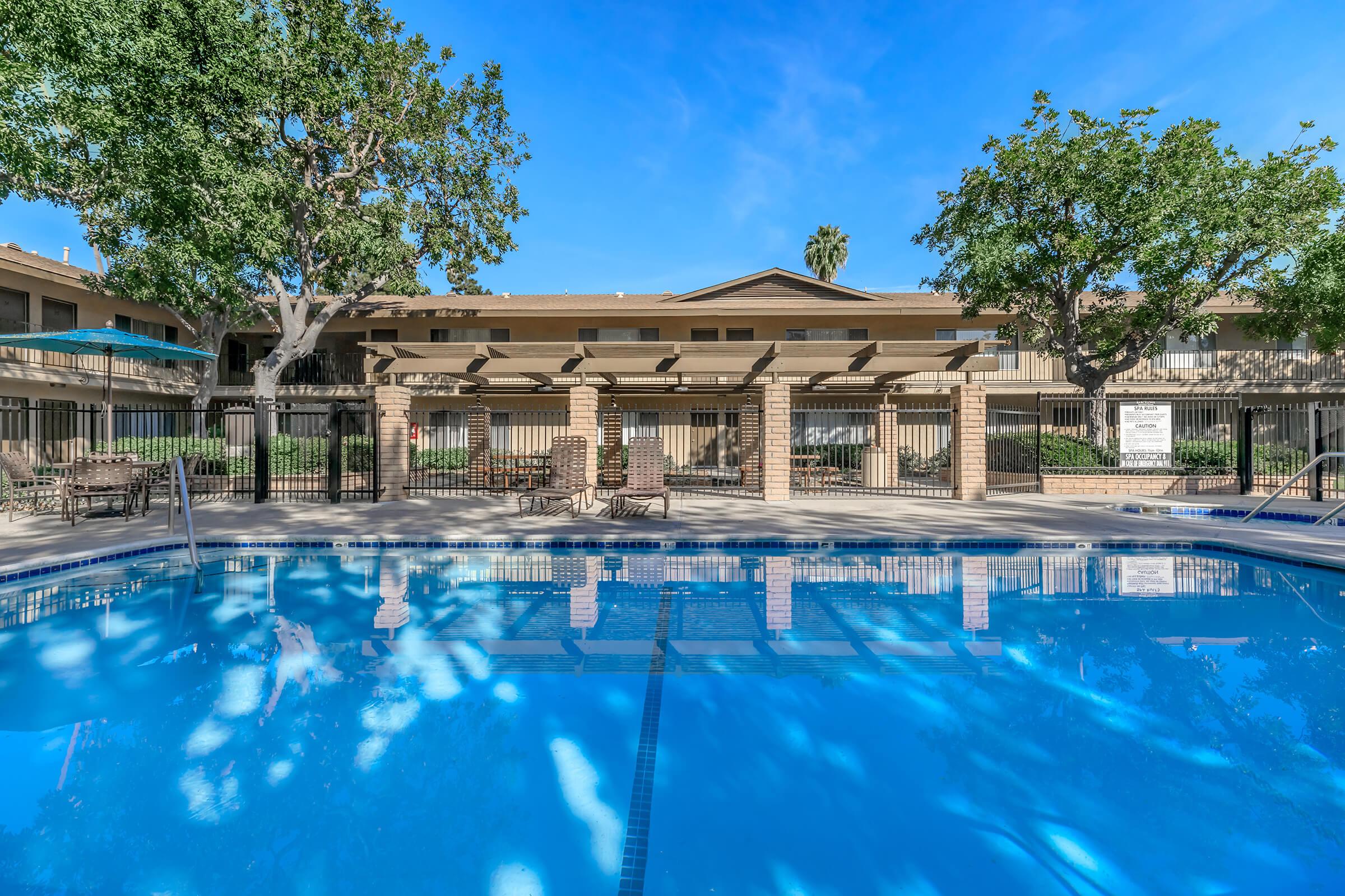 A clear blue swimming pool surrounded by lounge chairs and shaded by trees, with an apartment complex visible in the background. The sky is bright and sunny, creating a vibrant, inviting atmosphere. The area is well-maintained with a fence enclosing the pool.