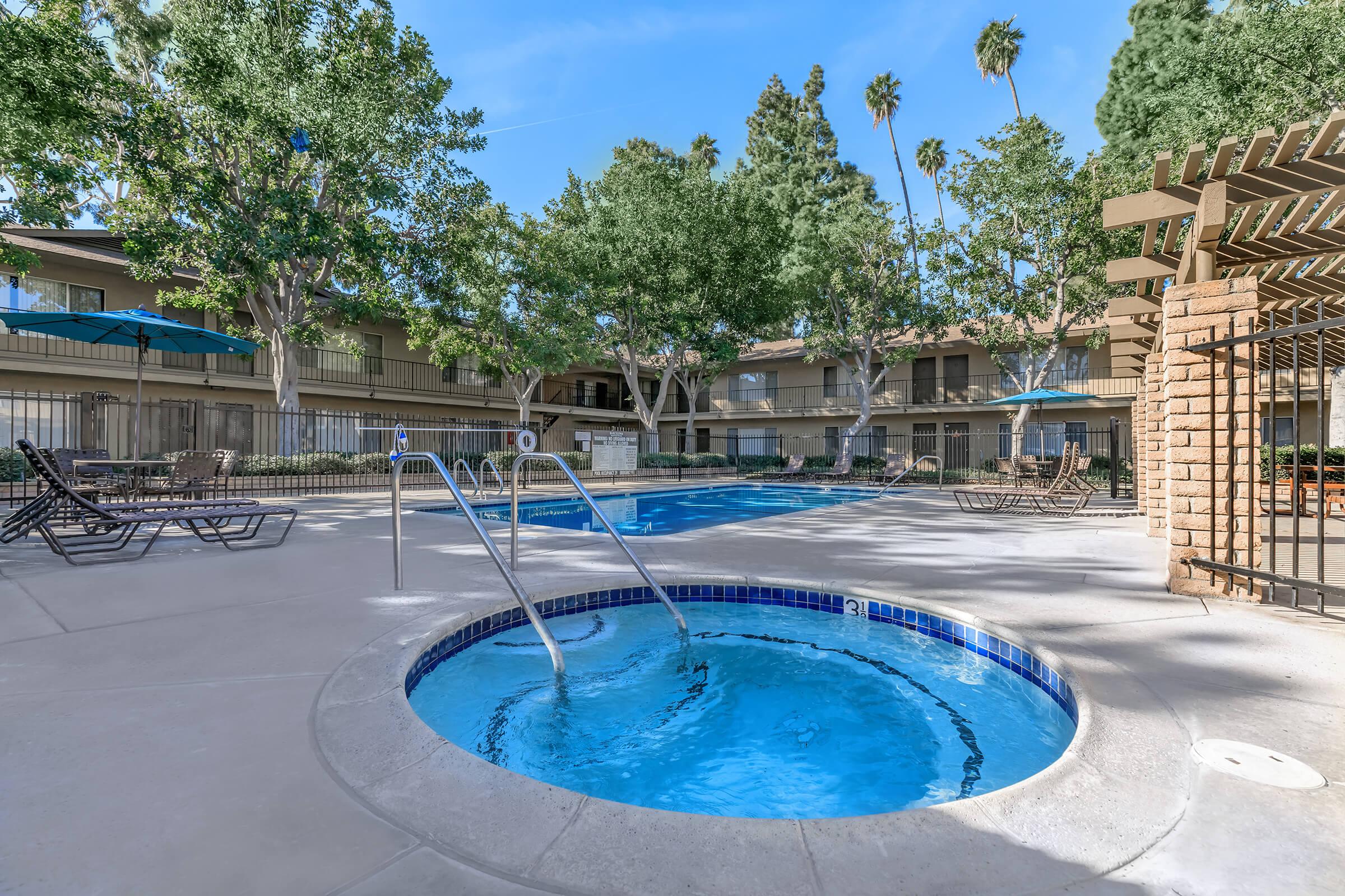 A serene pool area featuring a sparkling blue swimming pool and a hot tub. The space is surrounded by lush greenery and palm trees, with patio seating and umbrellas for shade, creating a relaxing outdoor environment for leisure and relaxation.