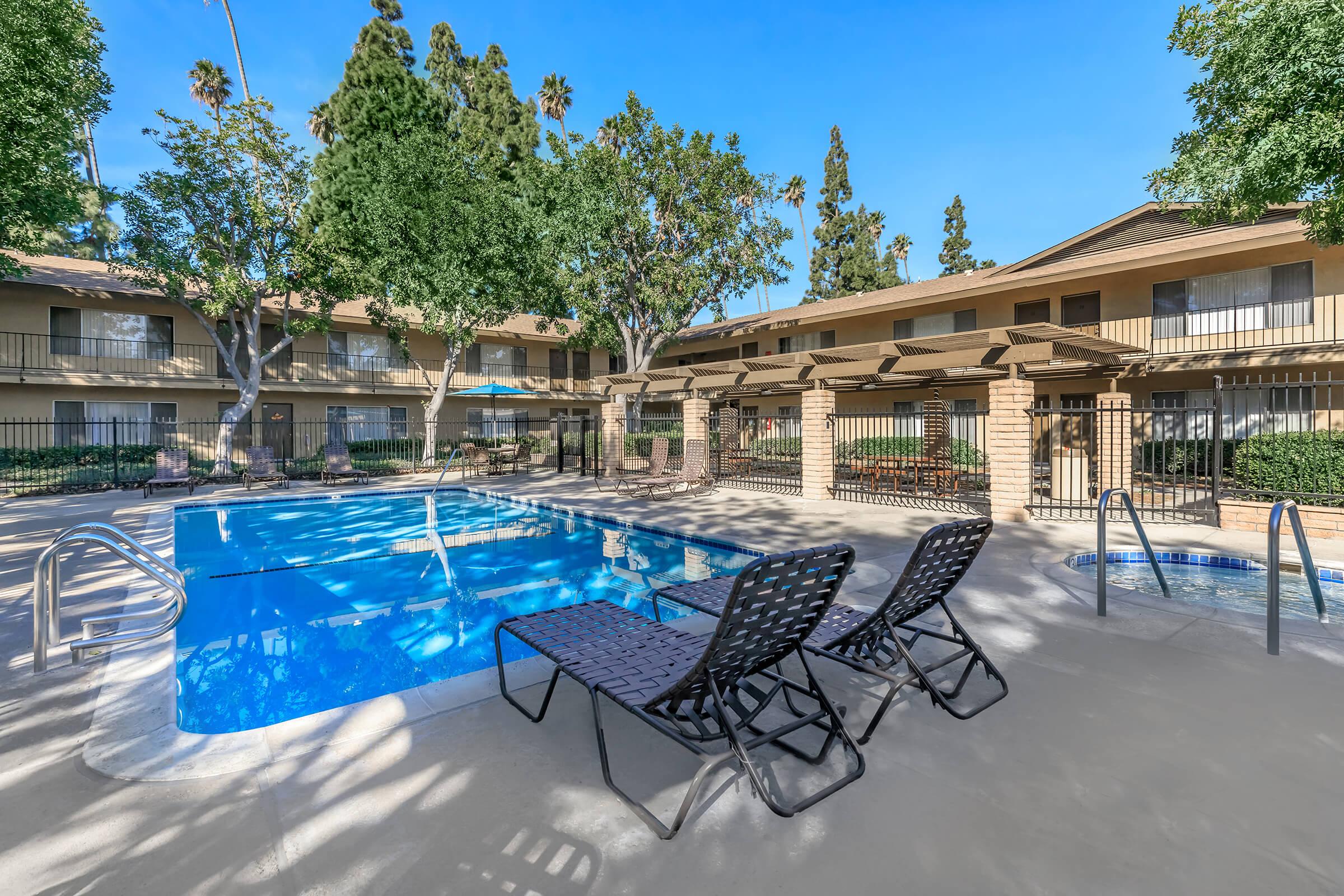 A clear blue swimming pool surrounded by lounge chairs, with trees and landscaped greenery in the background. Two building façades are visible in the background, each featuring balconies. The environment suggests a sunny, relaxing atmosphere typical of apartment complexes.