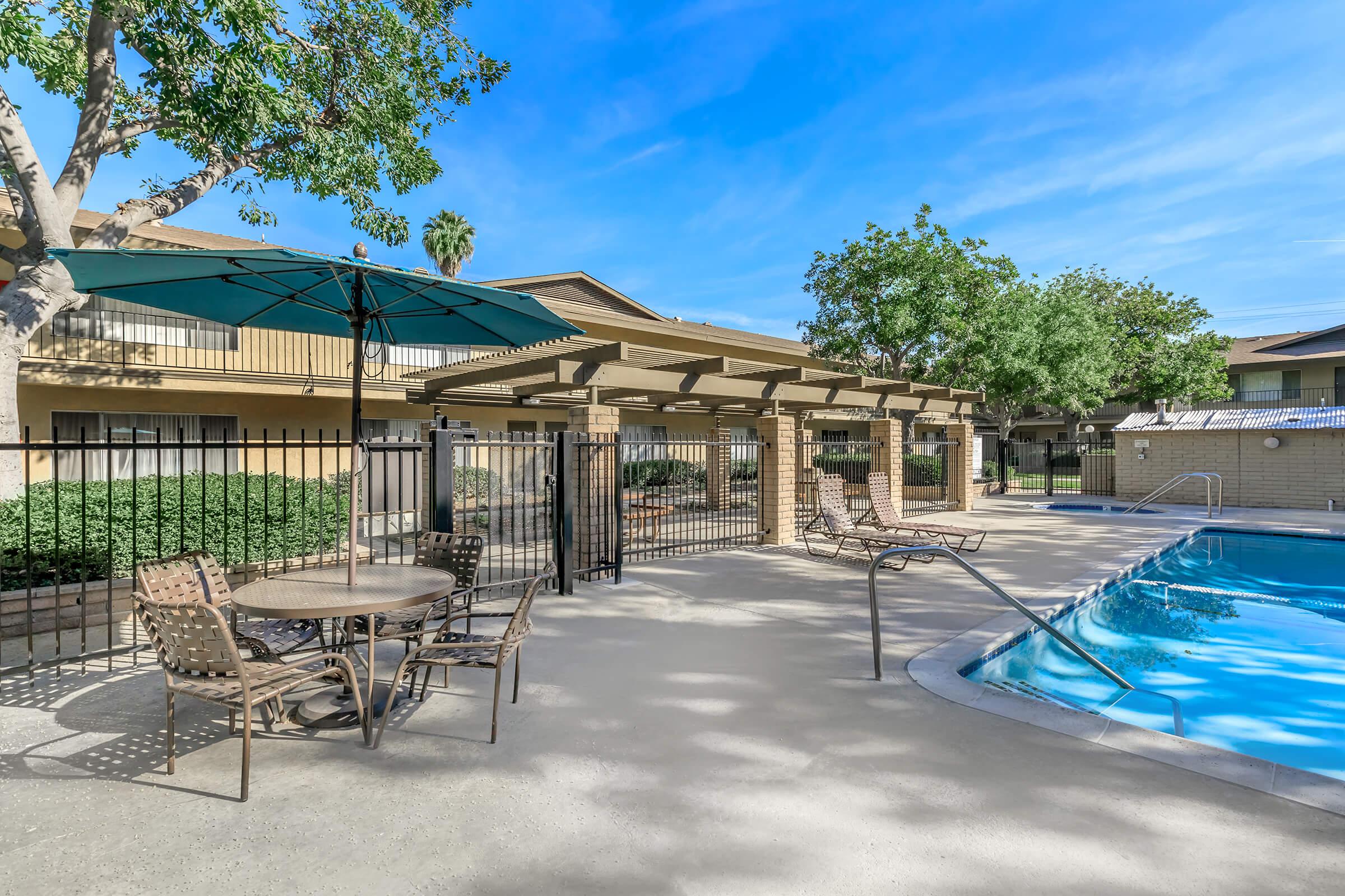 A pool area featuring a blue swimming pool, surrounded by a fenced patio with lounge chairs and a table under a green umbrella. Lush trees and landscaping provide shade, and residential buildings are visible in the background under a clear blue sky.