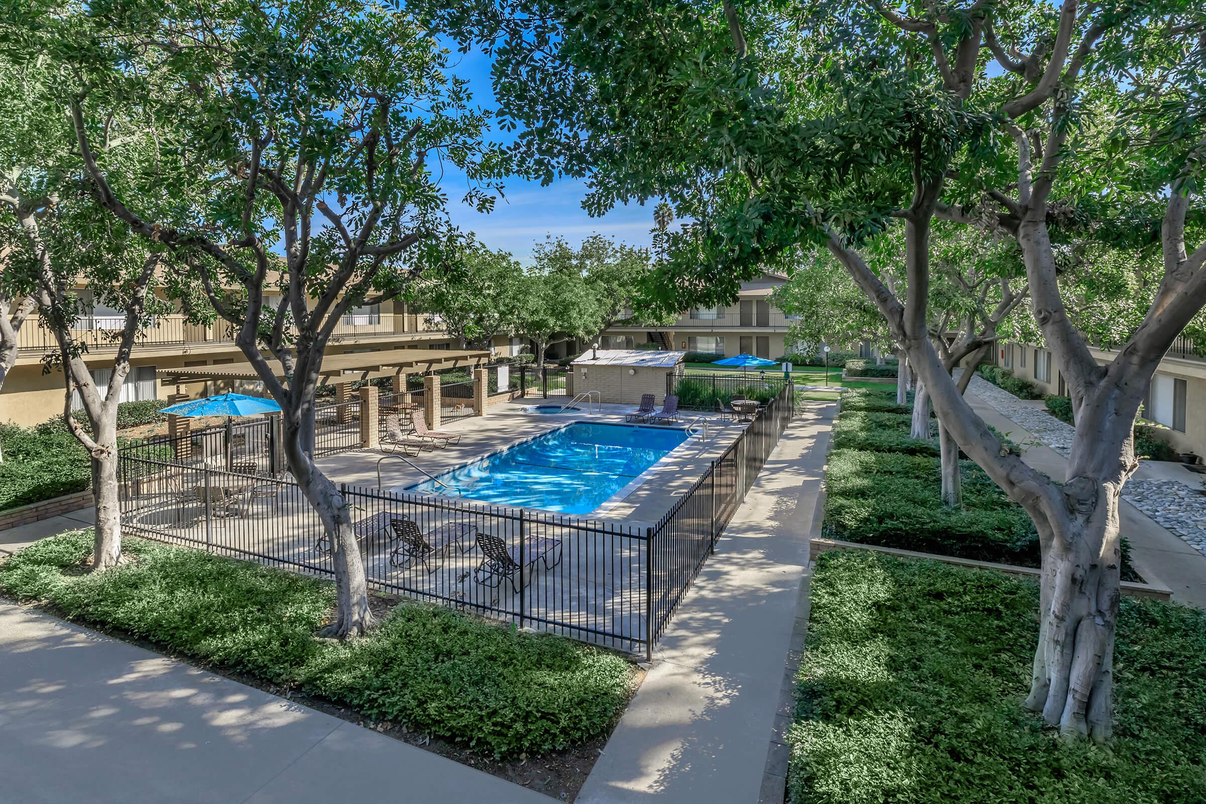 View of a well-maintained outdoor swimming pool surrounded by lush greenery and trees. The pool area is enclosed by a black fence and features lounge chairs and shaded umbrellas. Nearby, there are pathways connecting to residential buildings, creating a serene and inviting atmosphere.