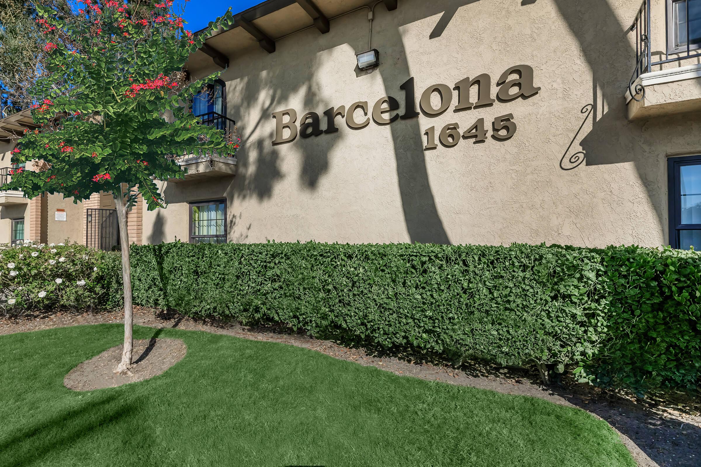 A building exterior with the word "Barcelona" and the number "1645" prominently displayed. The facade is beige with decorative elements. There's a small tree and well-maintained green shrubs in the foreground, complemented by a manicured lawn. The scene is bright with clear blue skies.