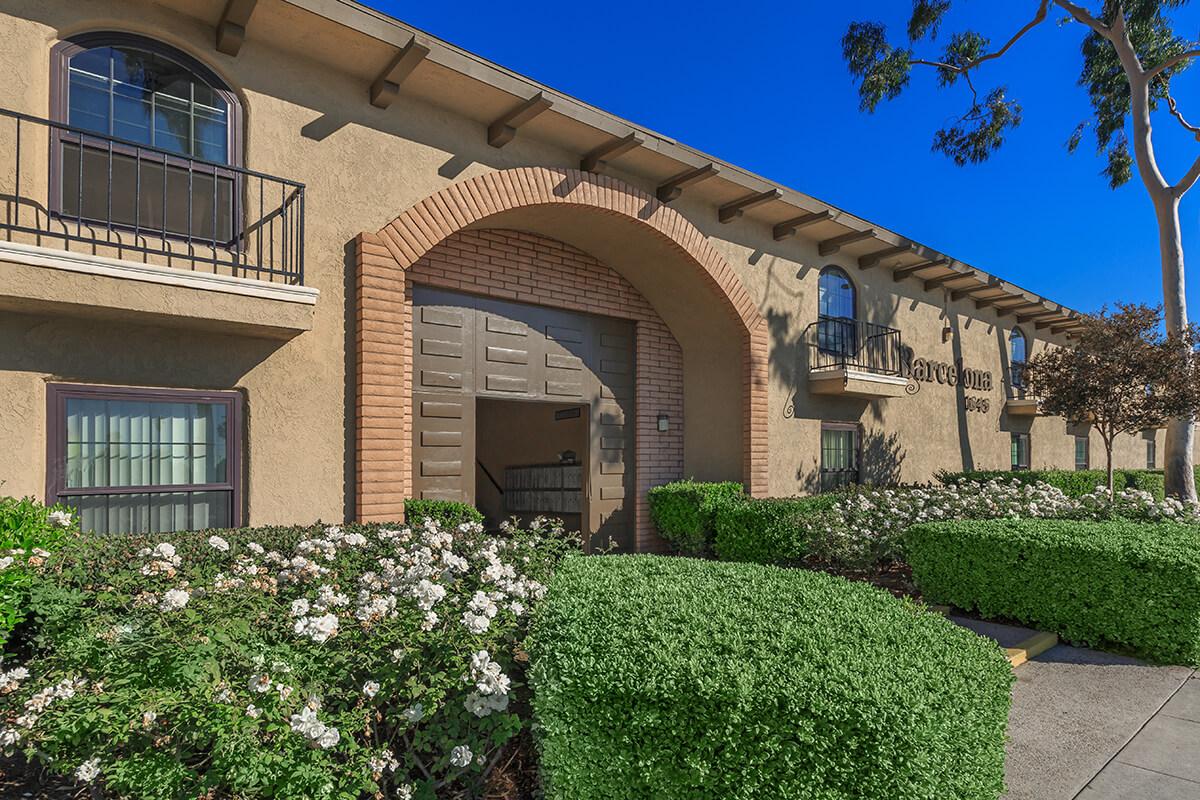 Exterior view of an apartment building featuring an arched entrance door, balconies with wrought iron railings, and landscaped greenery including hedges and flowering shrubs under a clear blue sky.