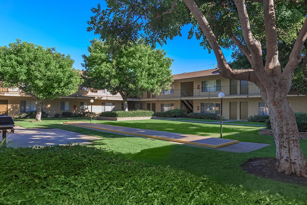 A well-maintained courtyard of an apartment complex, featuring lush green grass, several trees, and a walking path. The buildings have balconies and are set against a clear blue sky, creating a serene and inviting atmosphere.