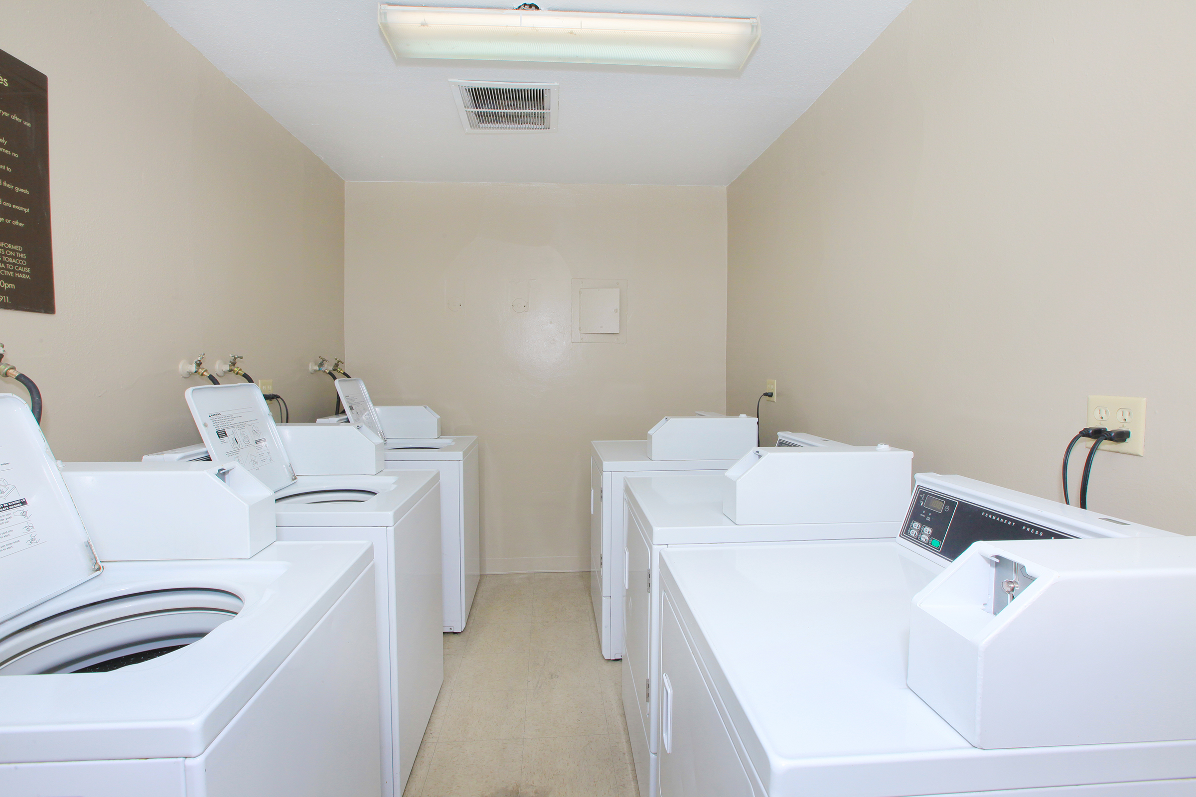 A clean, well-lit laundry room featuring several white washing machines aligned against the wall. The room has beige walls and a simple overhead light, creating a tidy atmosphere for washing clothes.