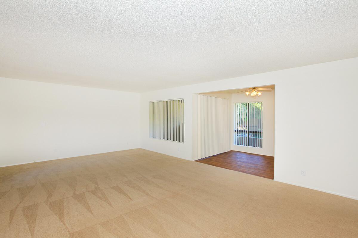 Spacious, empty living room with light beige carpet. White walls and large windows letting in natural light. A sliding glass door leads to an adjacent room, potentially a dining area or patio, separated by vertical blinds. The space has a bright and airy feel, perfect for personalization and decoration.